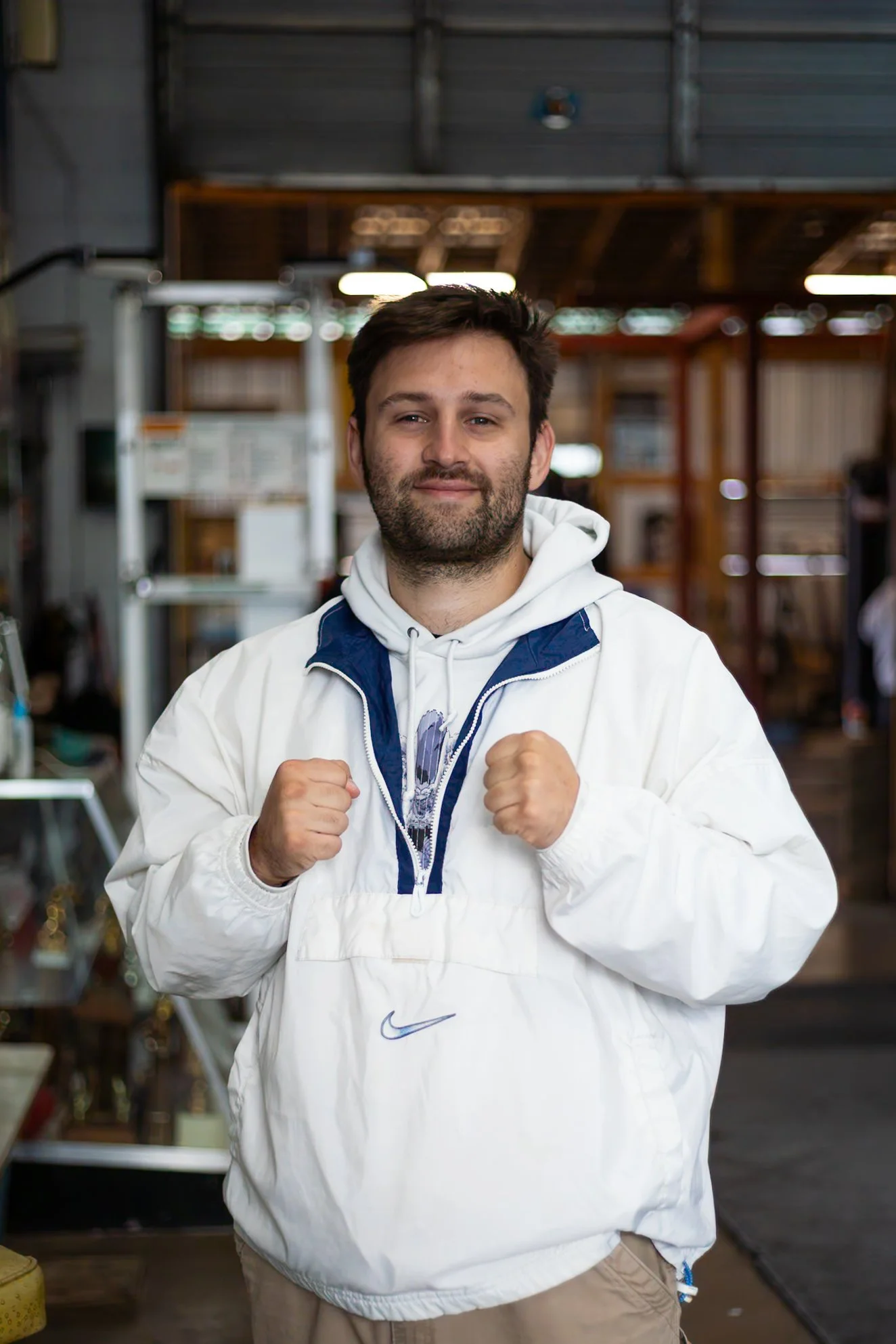 Young man with a beard dressed in a white Nike jacket and hoodie, fist clenched, smiling in a warehouse or industrial space with shelves and miscellaneous items in the background.