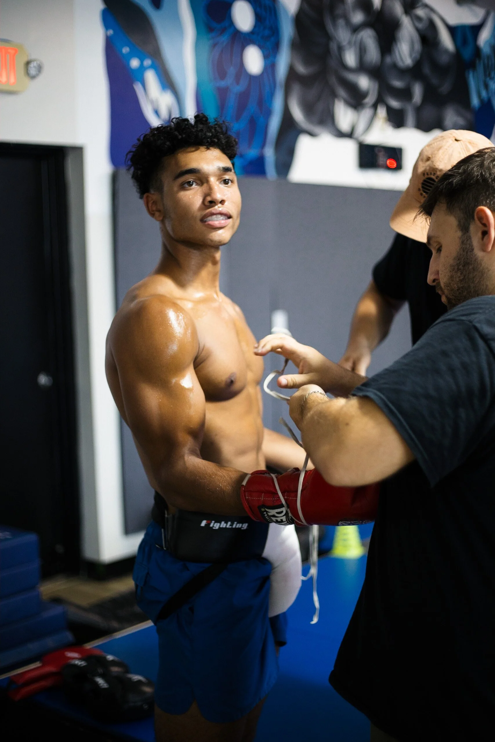 A male boxer with sweaty skin gets his boxing gloves adjusted by a team member inside a gym, with blue and black wall art in the background.