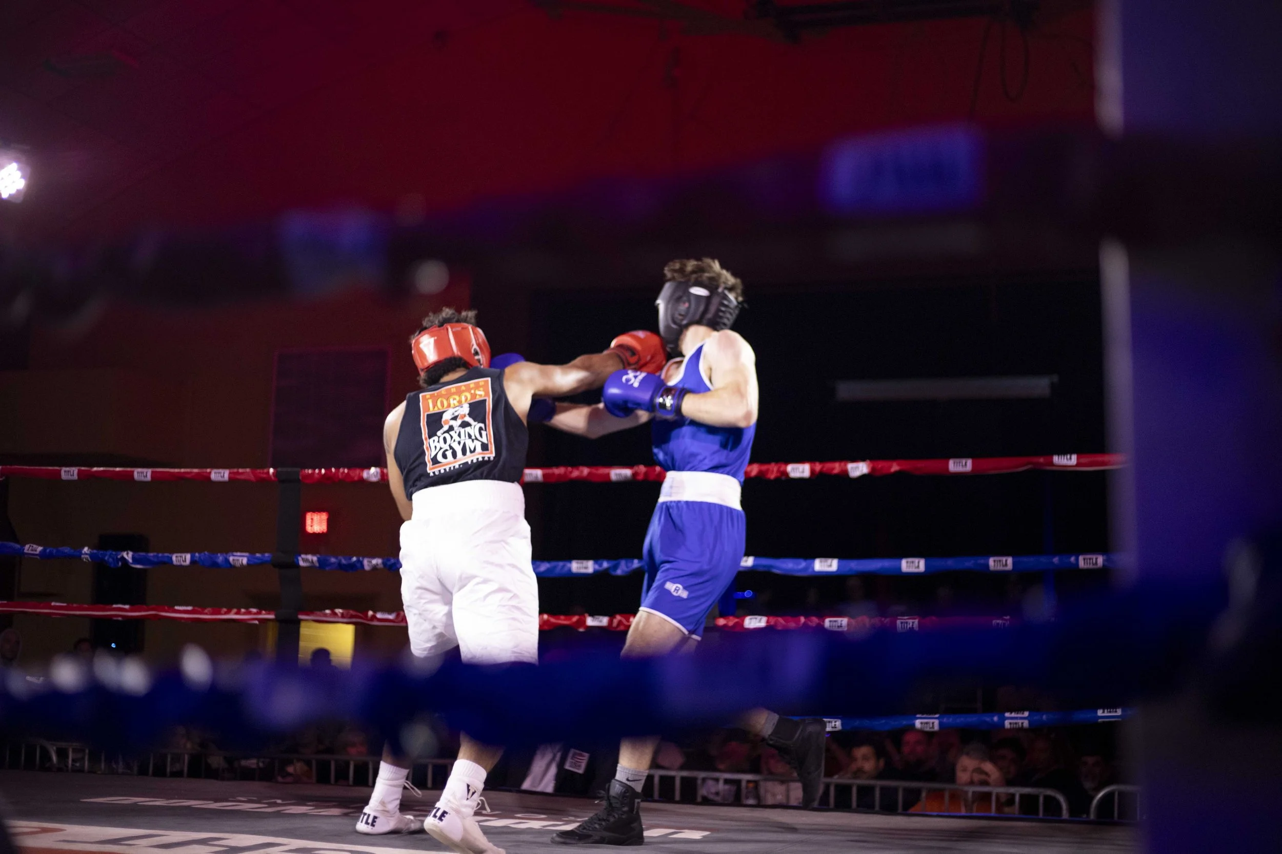 Two boxers in a ring engaged in a boxing match, with one delivering a punch to the other's face, both wearing protective headgear and gloves.