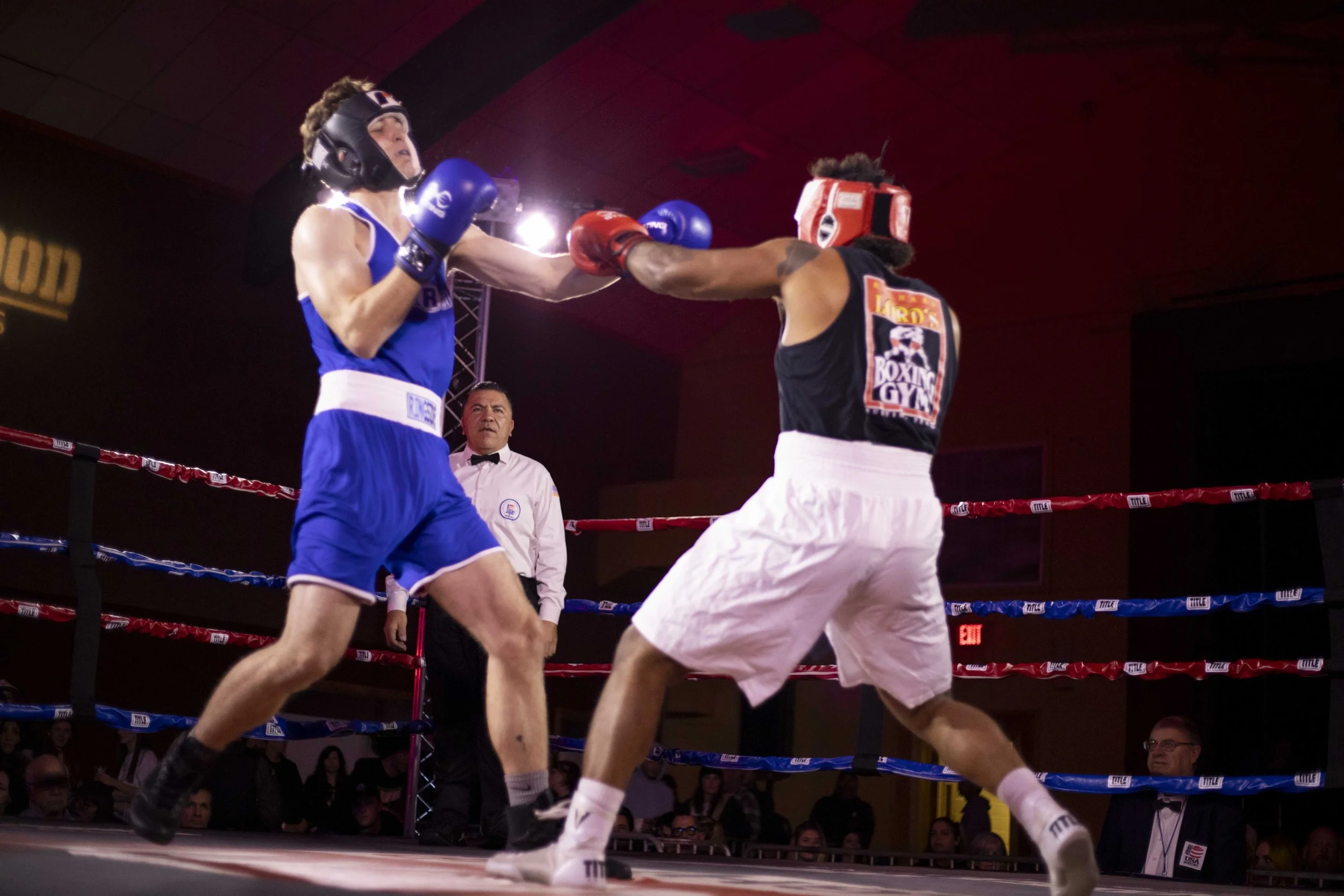 Two male boxers in a boxing ring throwing punches at each other, with a referee watching closely in the background, surrounded by spectators.