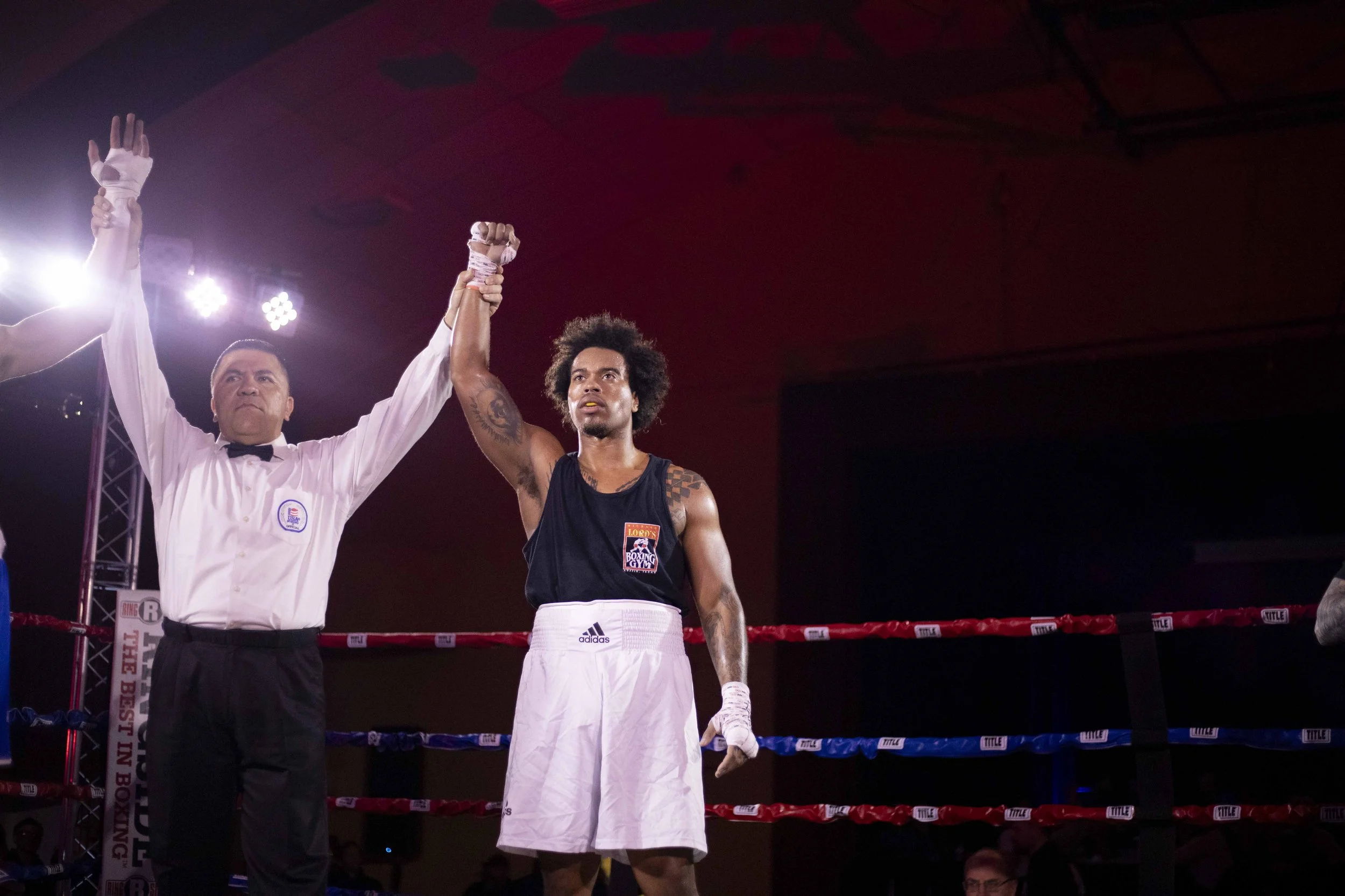A boxing match winner with curly hair wearing white shorts and a black tank top, standing in the boxing ring with a referee holding up his arm in victory.