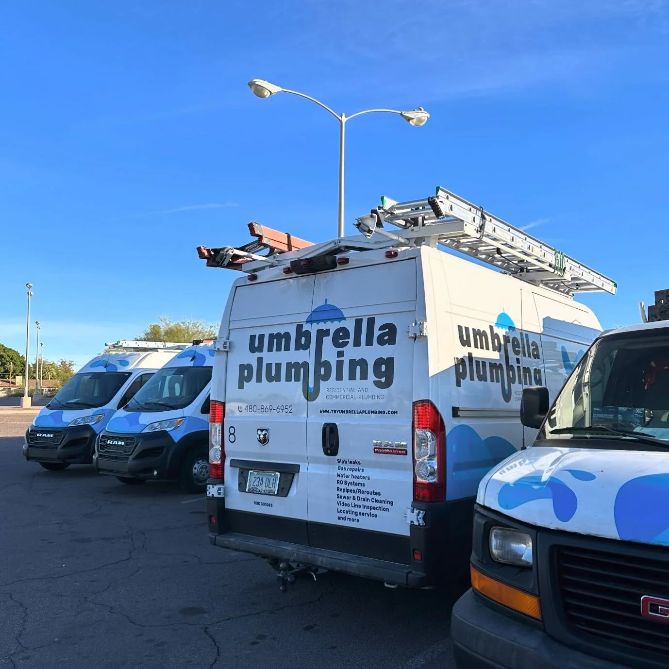 A white plumbing service van with the company name 'Umbrella Plumbing' and a blue umbrella logo, parked in a lot alongside two similar vans and a black GMC vehicle. The van has ladders and equipment on its roof.