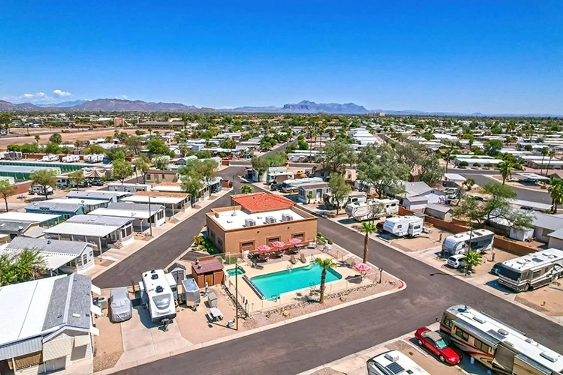 Aerial view of a desert mobile home park with a swimming pool, surrounded by RVs, palm trees, and grid-like streets, with distant mountains and a clear blue sky in the background.