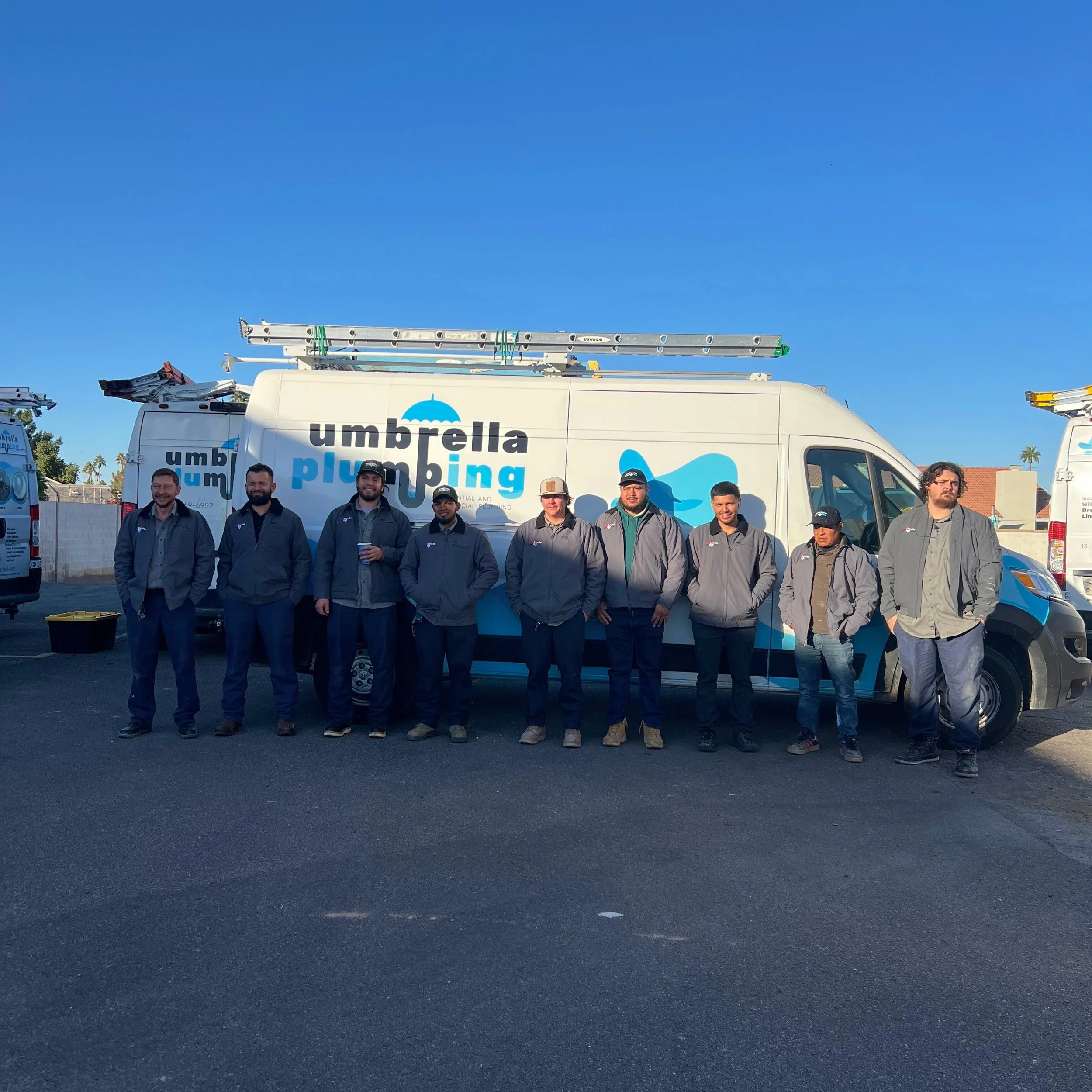 Group of nine people standing in front of a white plumbing service van with ladder on top, all wearing grey jackets and casual pants, in an outdoor parking lot.