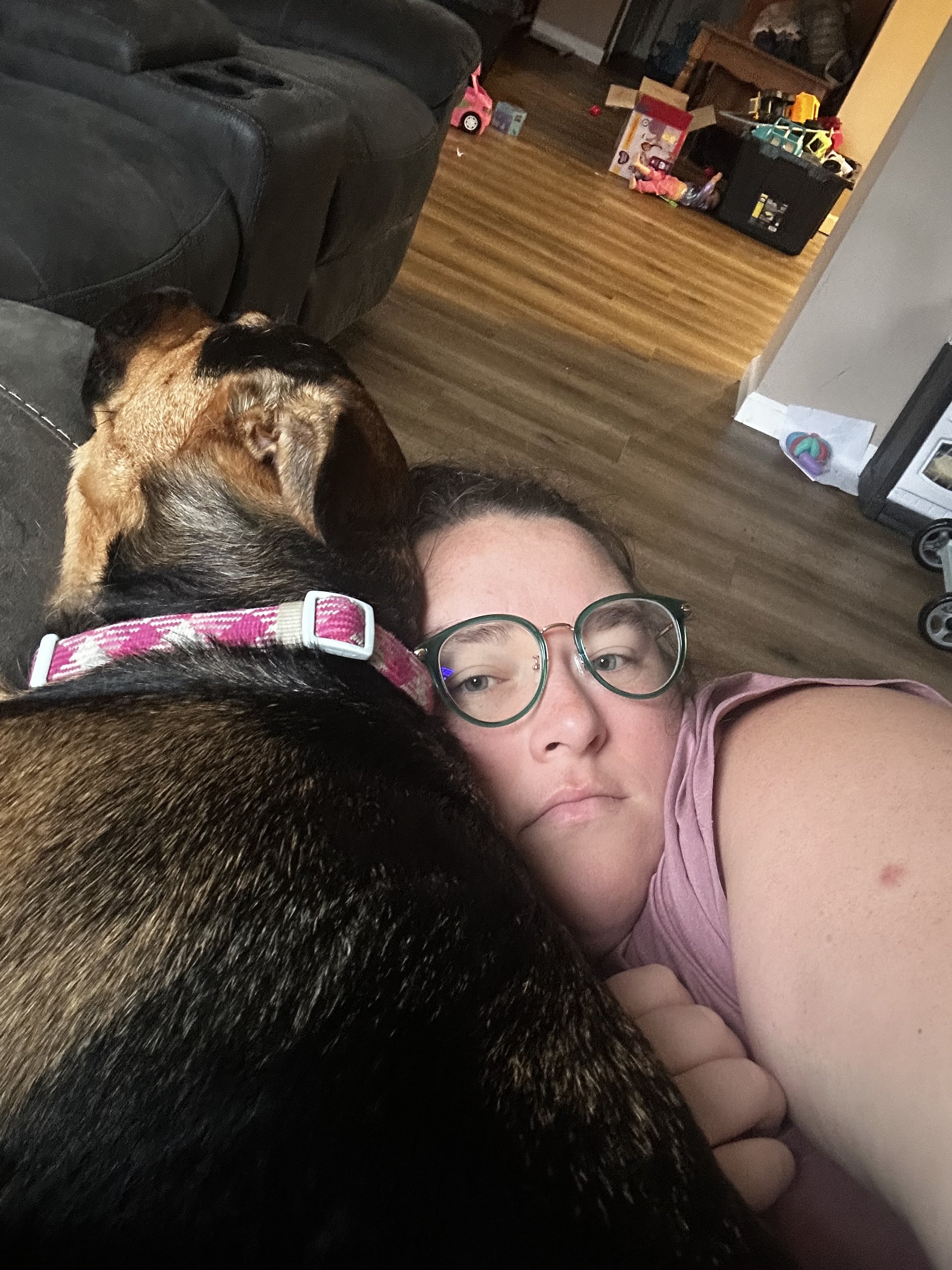 Person wearing glasses, lying on couch with a dog, in a living room with toys and boxes in the background.