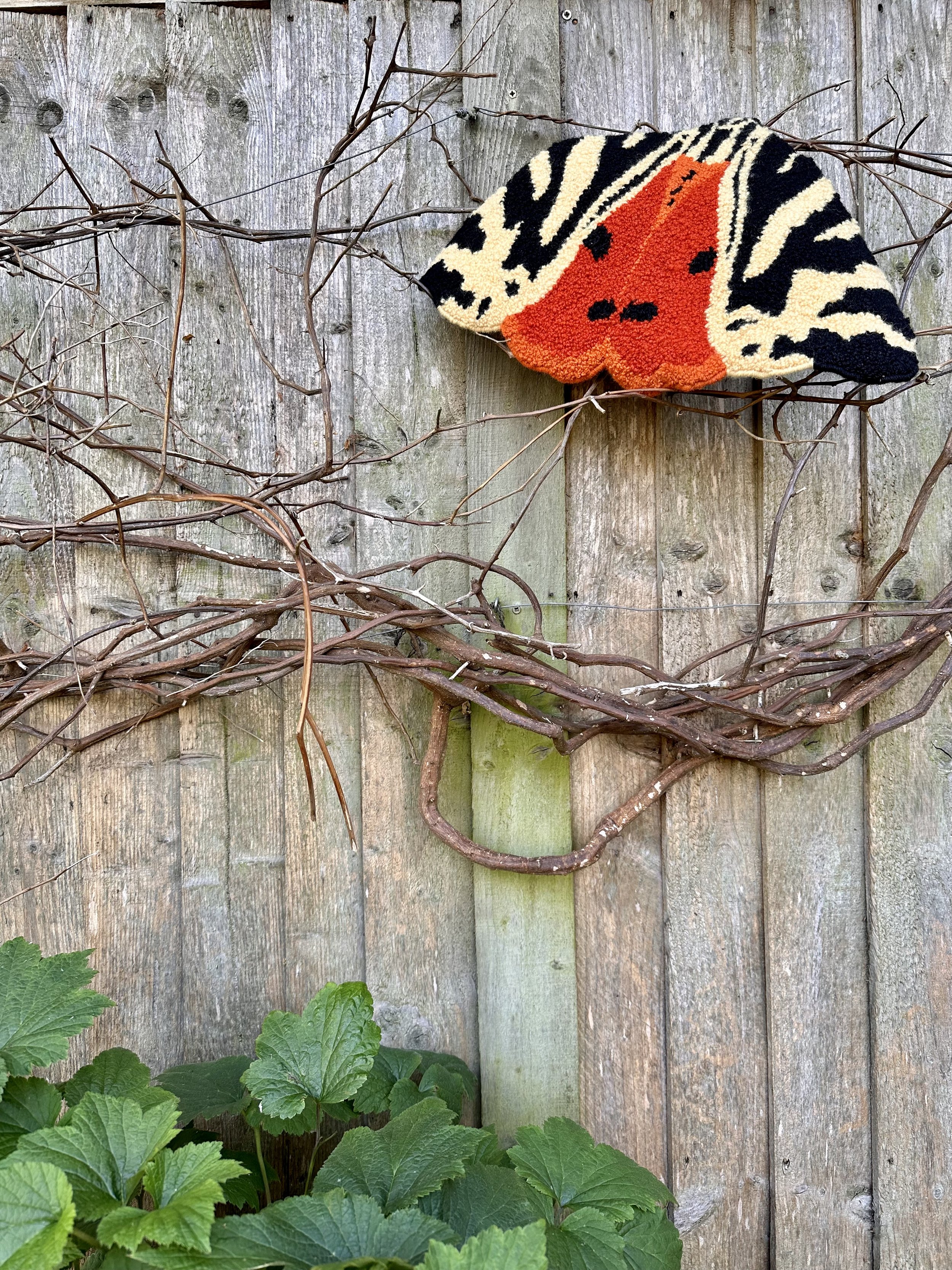 A multicolored knitted or woven fabric piece resembling a butterfly mounted on a wooden fence with bare vine branches draped around it, and green plants at the bottom.