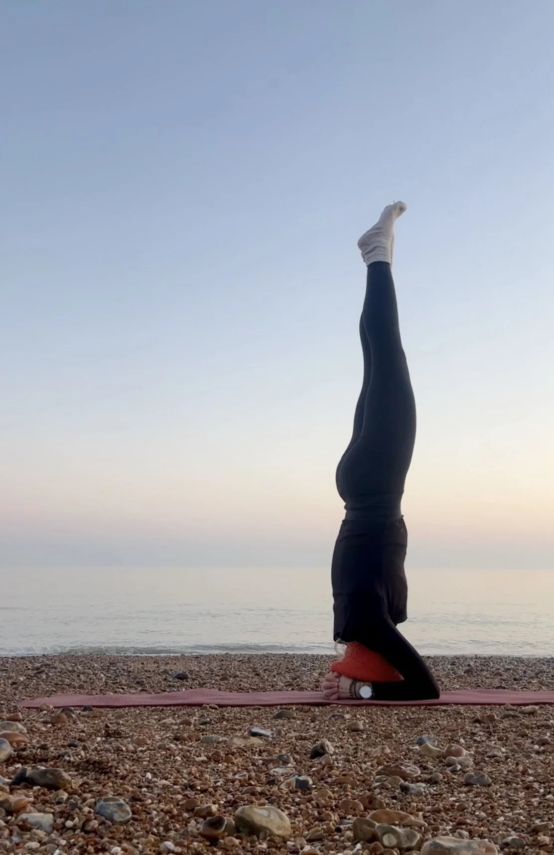 Person practicing a headstand yoga pose on a beach during sunrise or sunset.