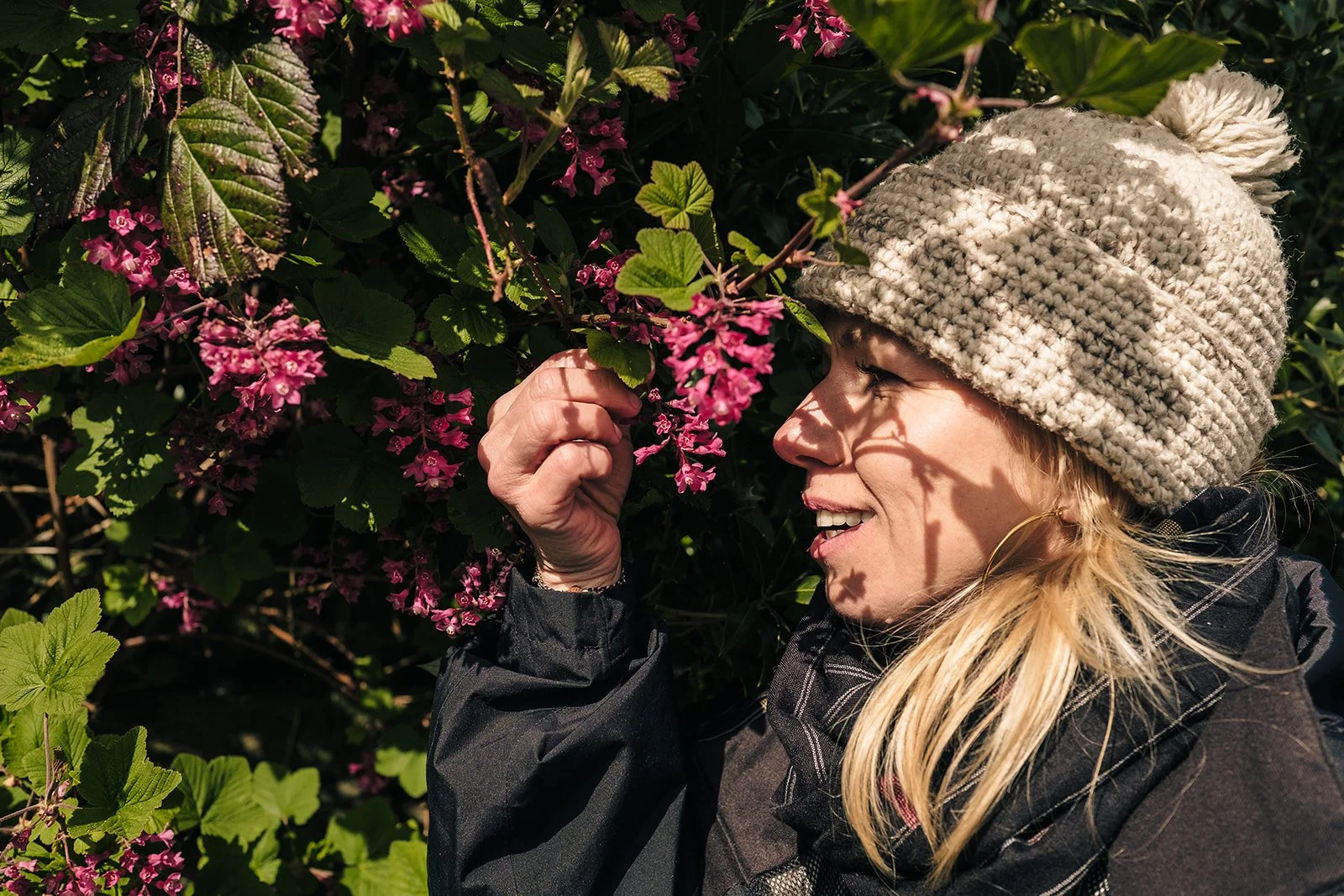 Person wearing a beige knit hat and scarf looks at pink flowers in a garden.