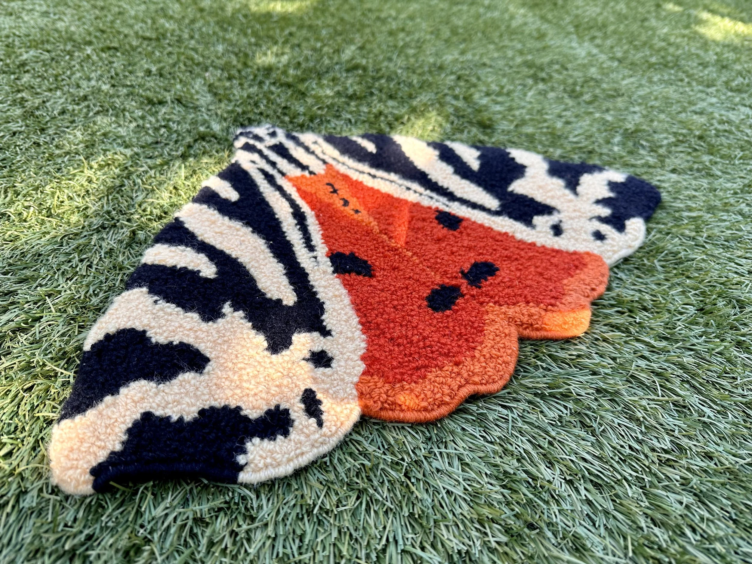 Colorful butterfly-shaped rug with black, orange, and cream patterns on a grass surface.