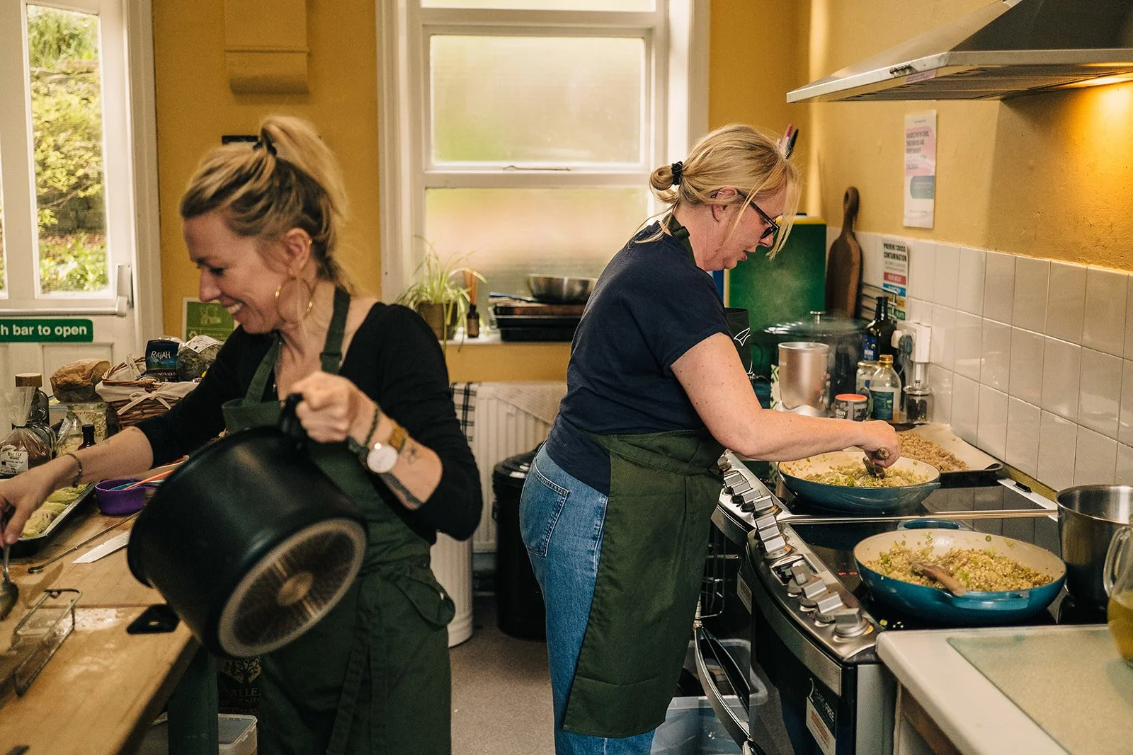 Two women cooking in a kitchen, both wearing green aprons. They are preparing food on a stovetop with pots and pans. The kitchen has yellow walls and a window in the background.