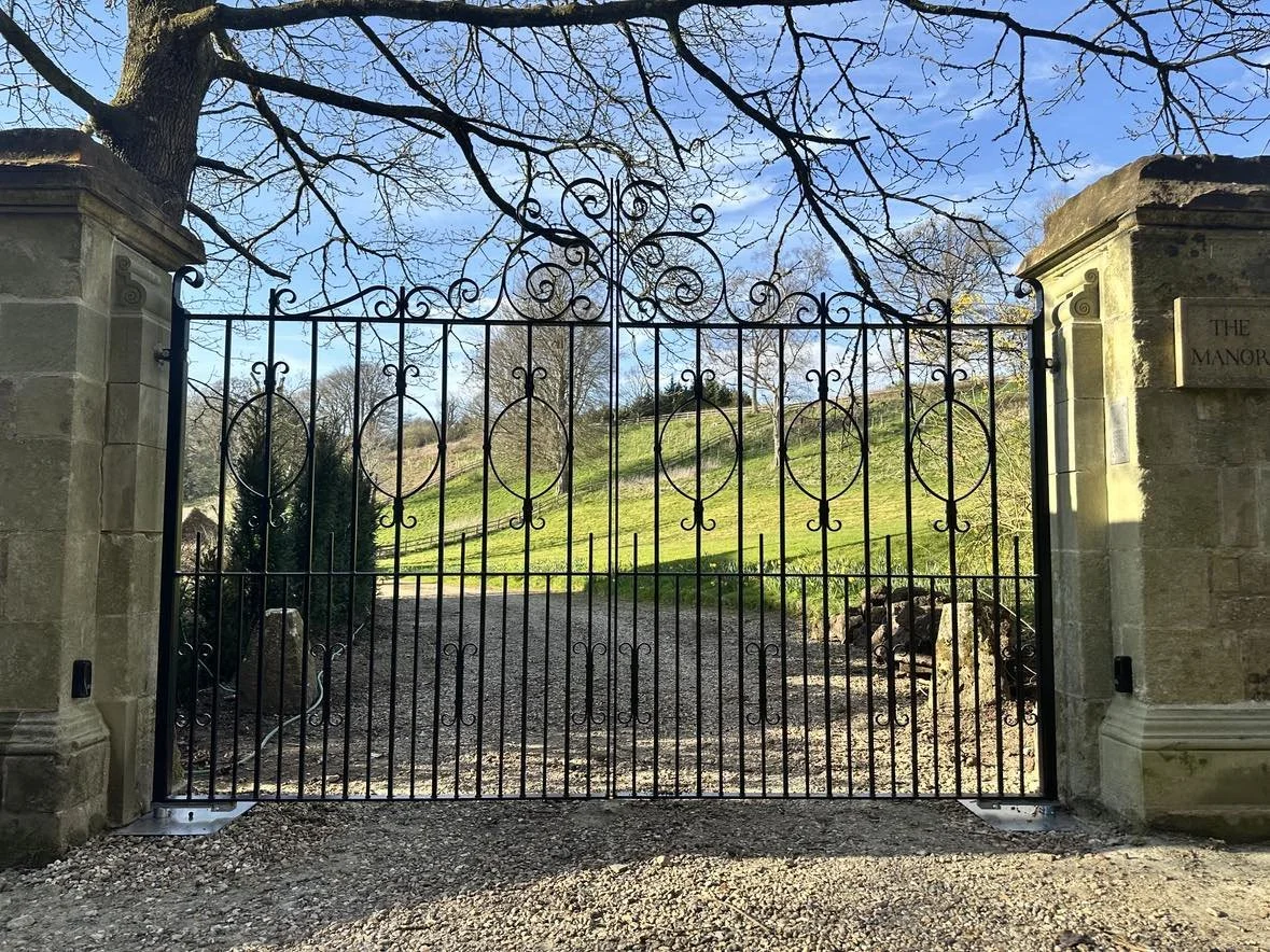 Gray double gate with wooden fence on both sides, set in yard with gravel ground, behind tall trees with leafless branches.