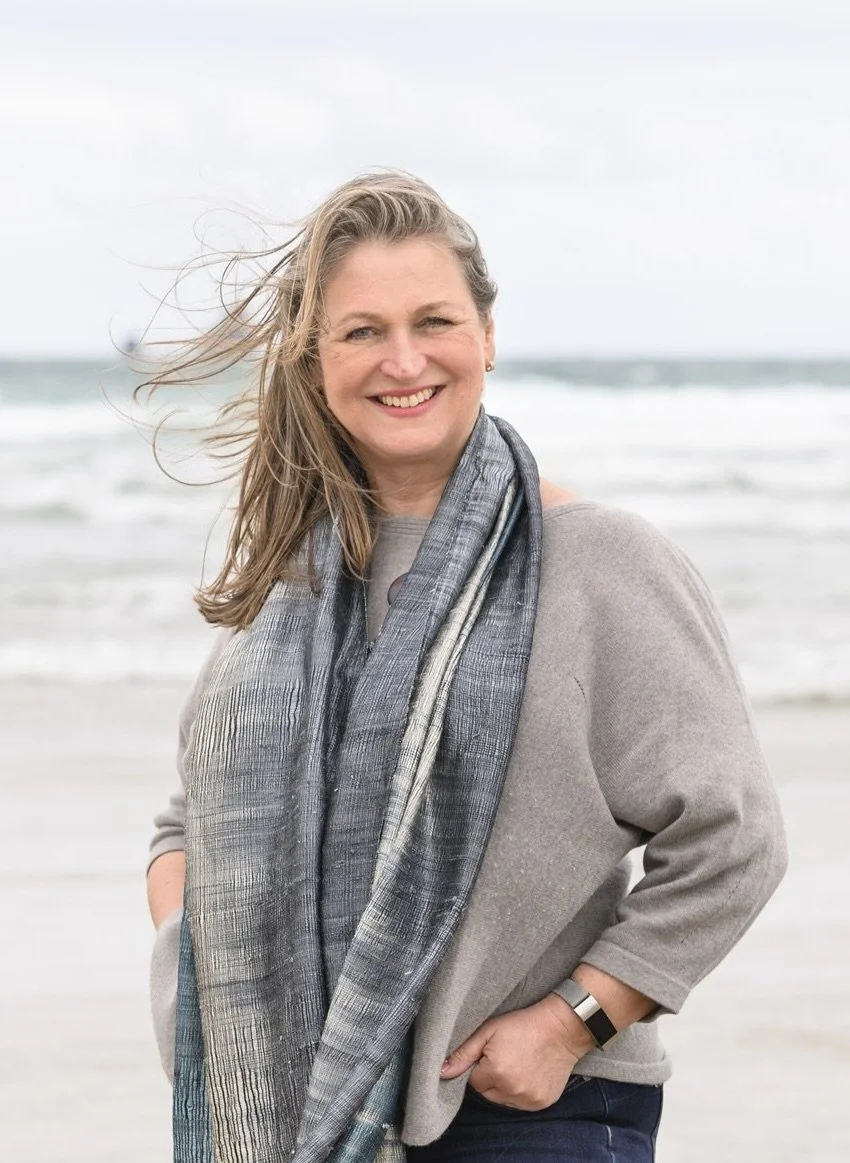 A woman with blonde hair smiling at the camera, standing on a beach with waves in the background, wearing a gray sweater and a gray scarf.