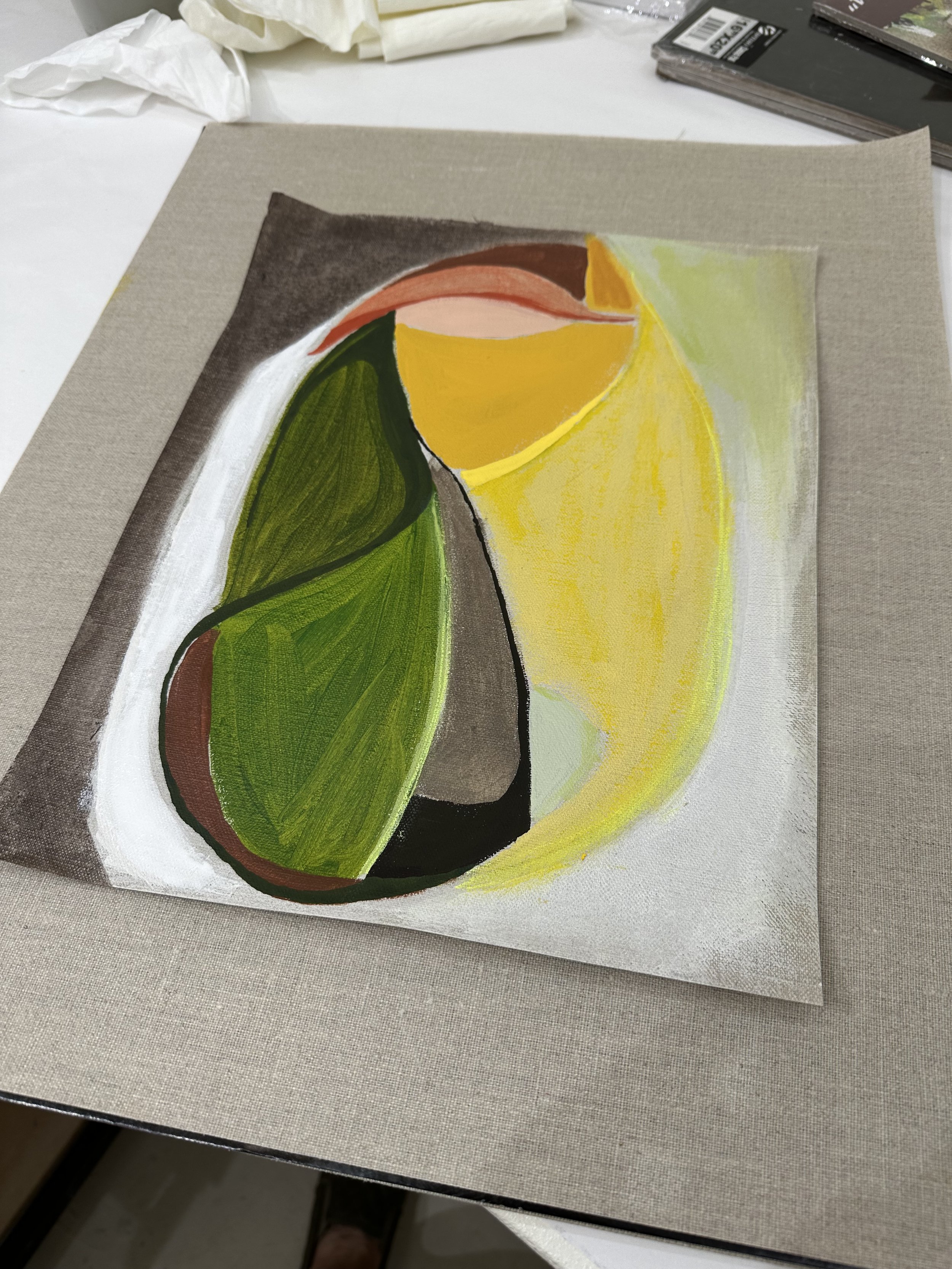 Abstract painting with green, yellow, brown, and peach shapes on canvas, lying on a table with art supplies.