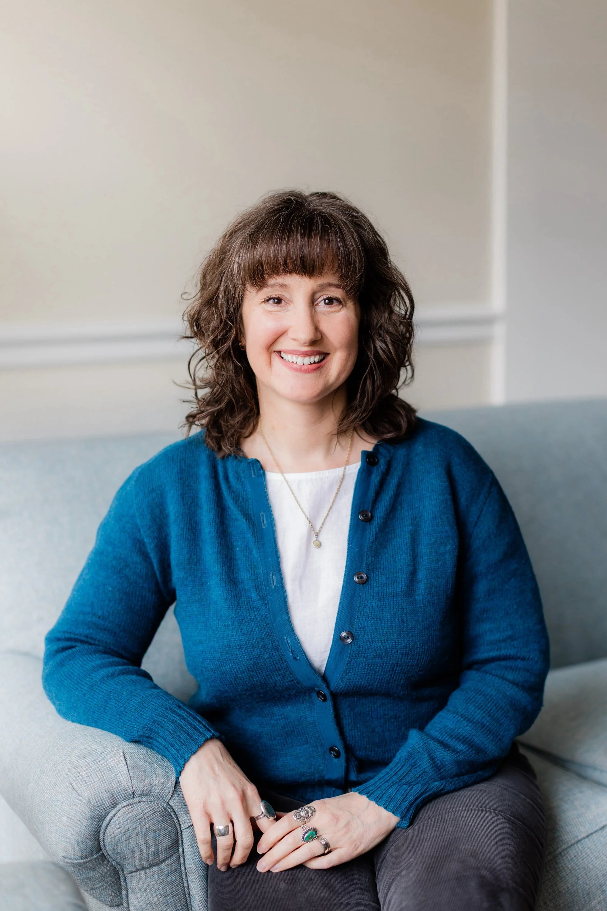 A woman with brown, curly hair smiling while sitting on a light blue sofa in a room with neutral walls, wearing a blue cardigan over a white top and multiple rings.