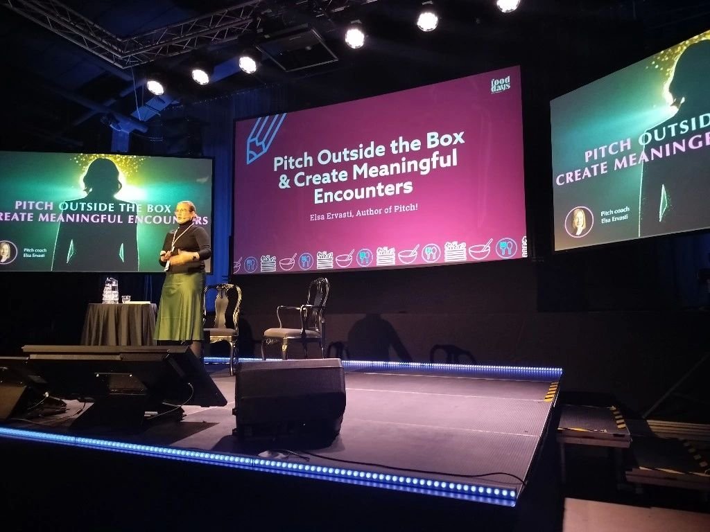 Kuvassa on nainen puhujapaikalla lavalla, jossa on kolme tuolia, ja kaksi suurta näyttöruutua, jotka esittävät samaa sisältöä: 'Pitch Outside the Box & Create Meaningful Encounters' sekä 'Elsa Ervasti, Author of Pitch!'.
