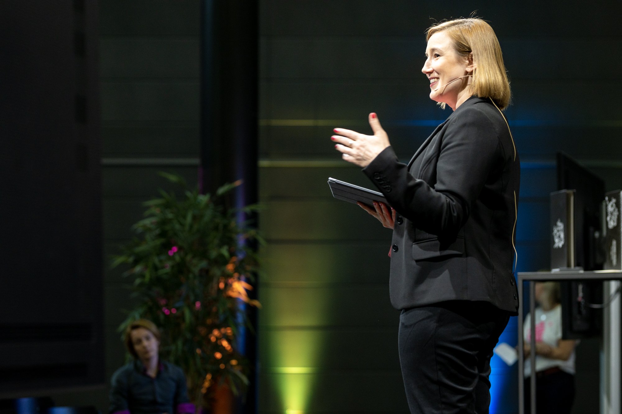 Nainen puhuu lavalla, jossa on mikrofonikaapeli ja tabletti, reunustettu vihreillä ja sinisillä valoilla, taustalla kaksi ihmistä ja viherkasvi.