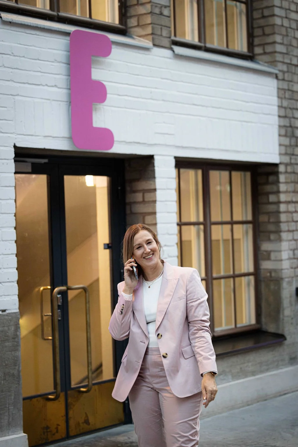 Naistapeili puhelimessa kävelee kadulla, taustalla rakennus ja suurin E-kirjaimin varustettu kyltti.