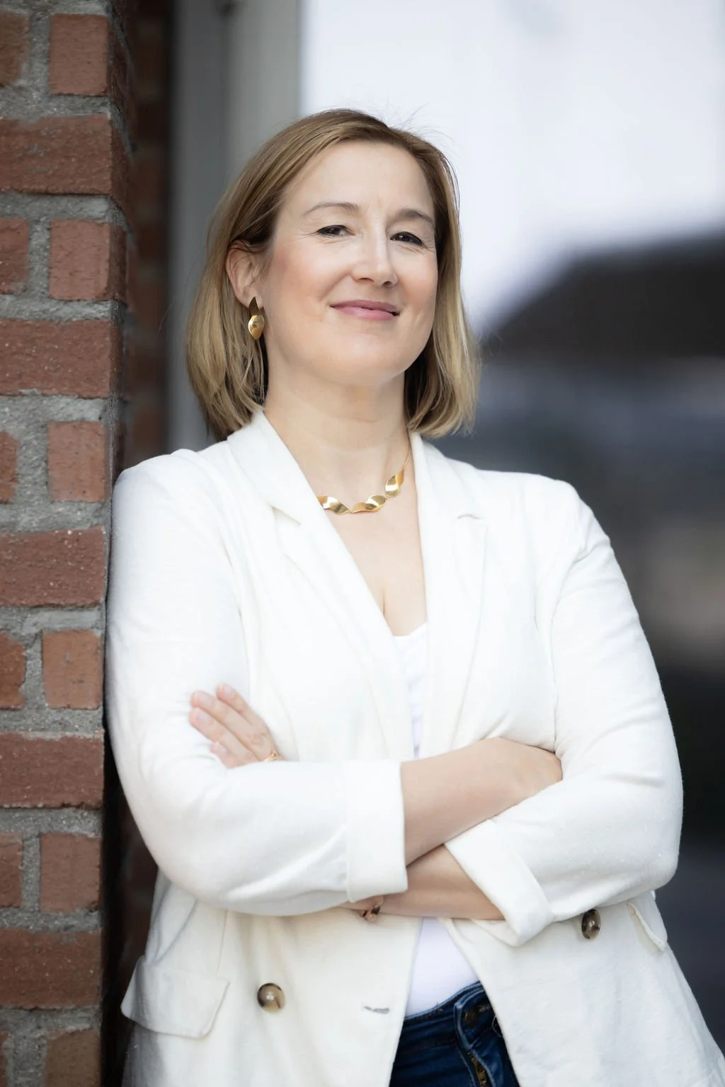 Nainen hymyilee ja seisoo ristissä käsivarsin vieri seinää vasten liikemiesvaatteissa, taustalla ulkoilma ja rakennus.