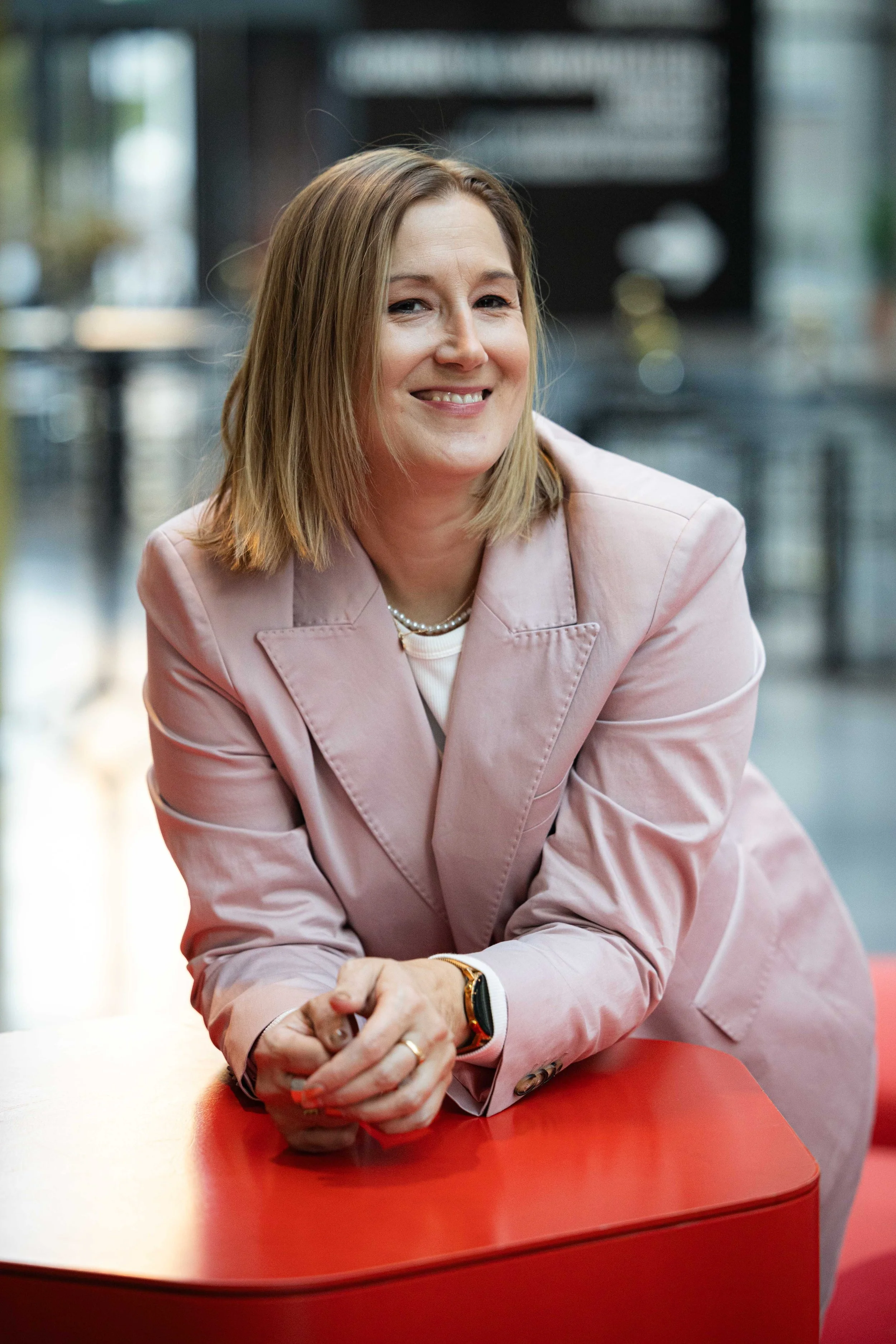 Nainen hymyilee ja istuu punaisella pöydällä, blair a pinki takki ja koruja, taustassa moderni sisätila.