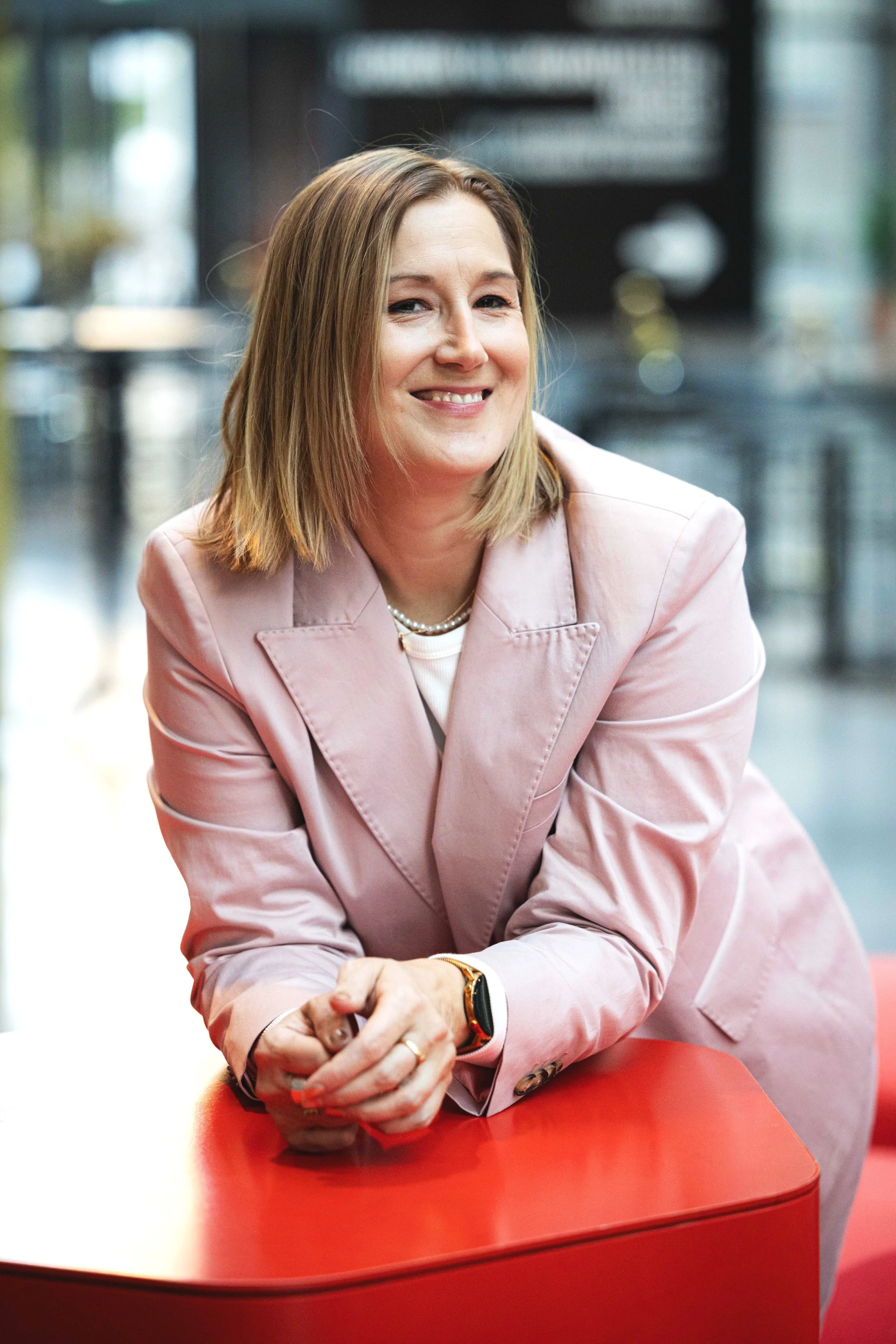 Nainen hymyilee ja istuu punaisella pöydällä, taustalla moderni sisätila, jossa on lasi- ja metallirakenteita.