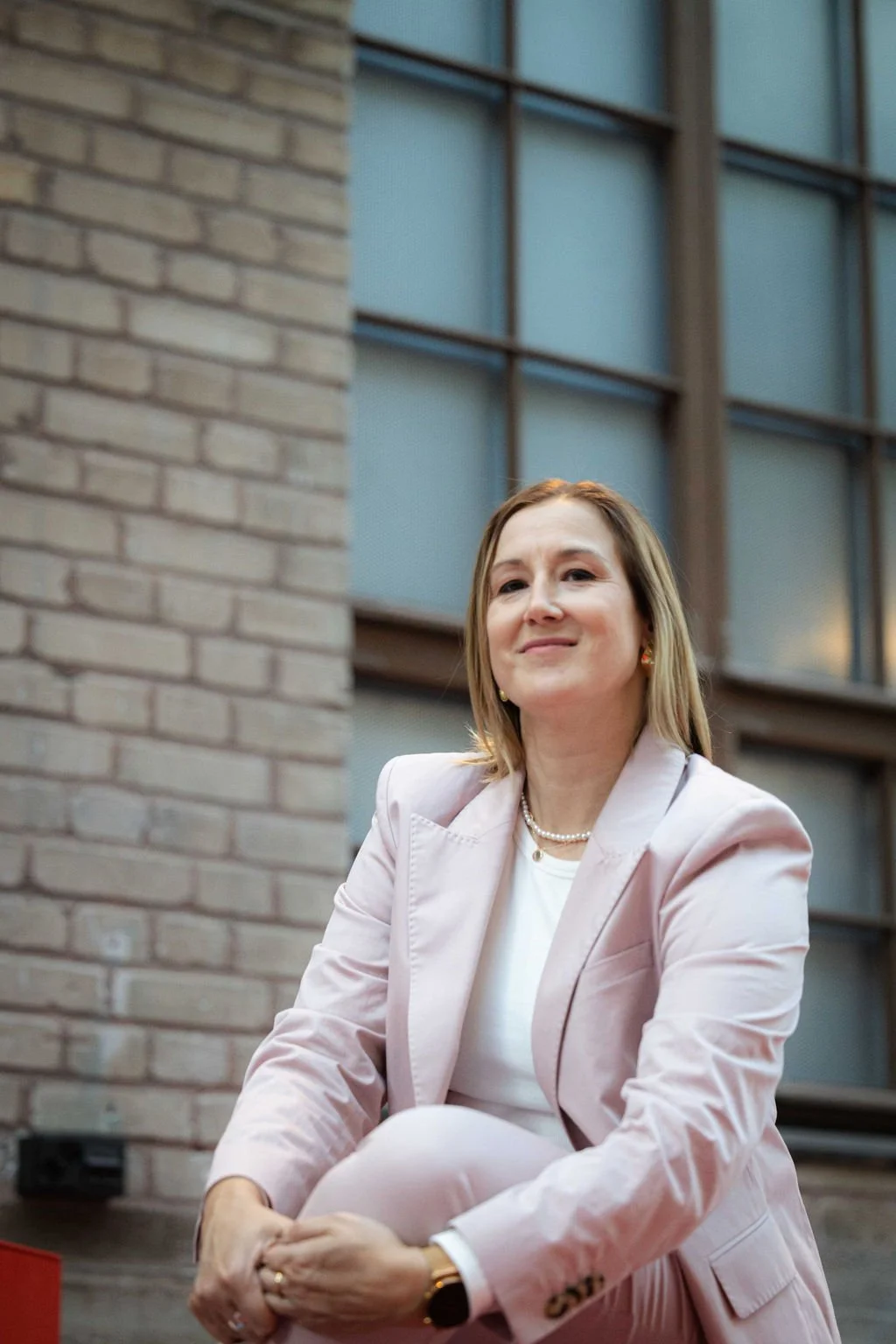 Nainen istuu ulkona kaupungin rakennuksen edessä, taustalla rakennuksen ikkunat ja muurirakennelma.