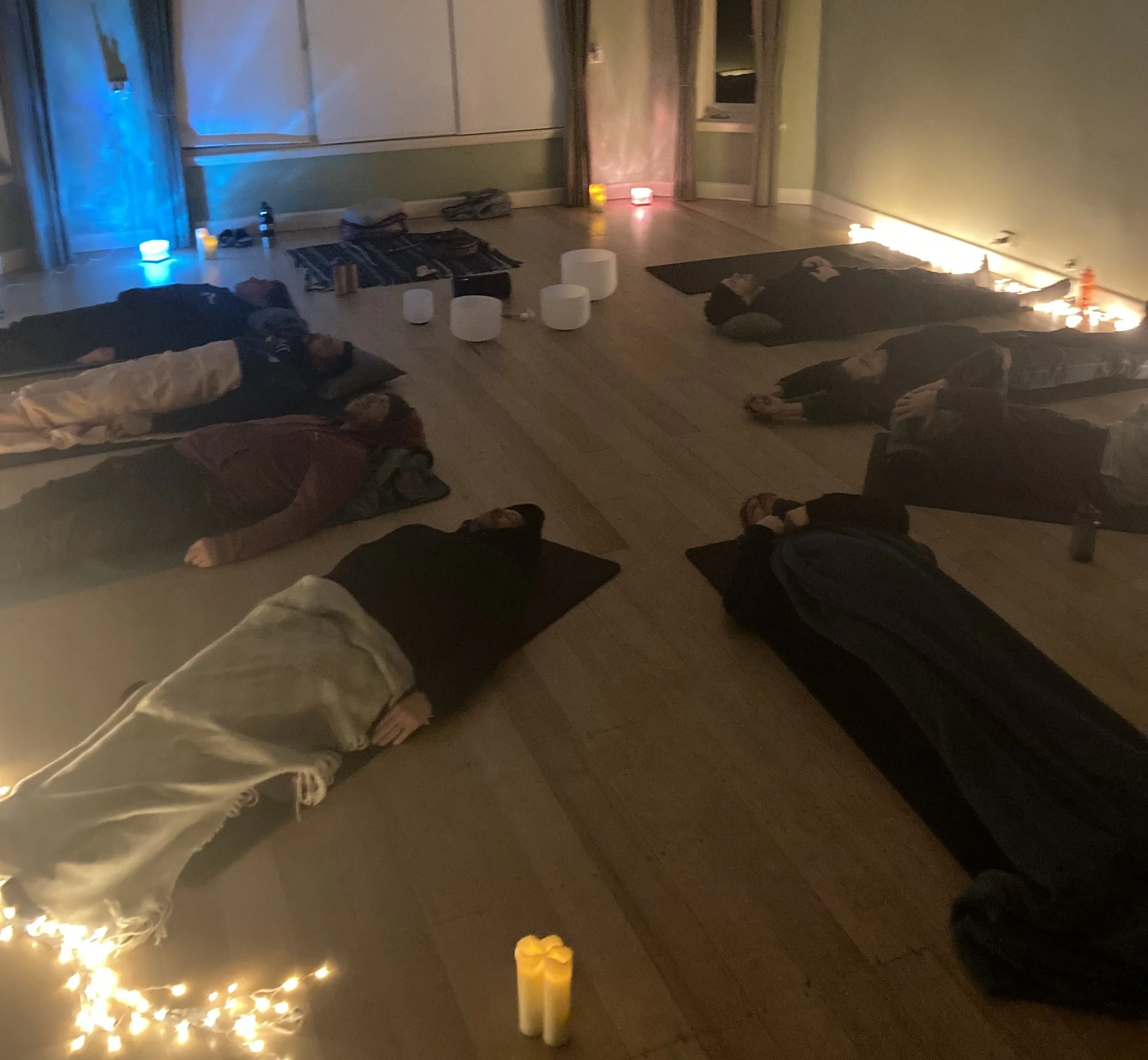 People lying on yoga mats in a candle lit room, participating in a meditation or relaxation session, surrounded by candles and soft lighting.