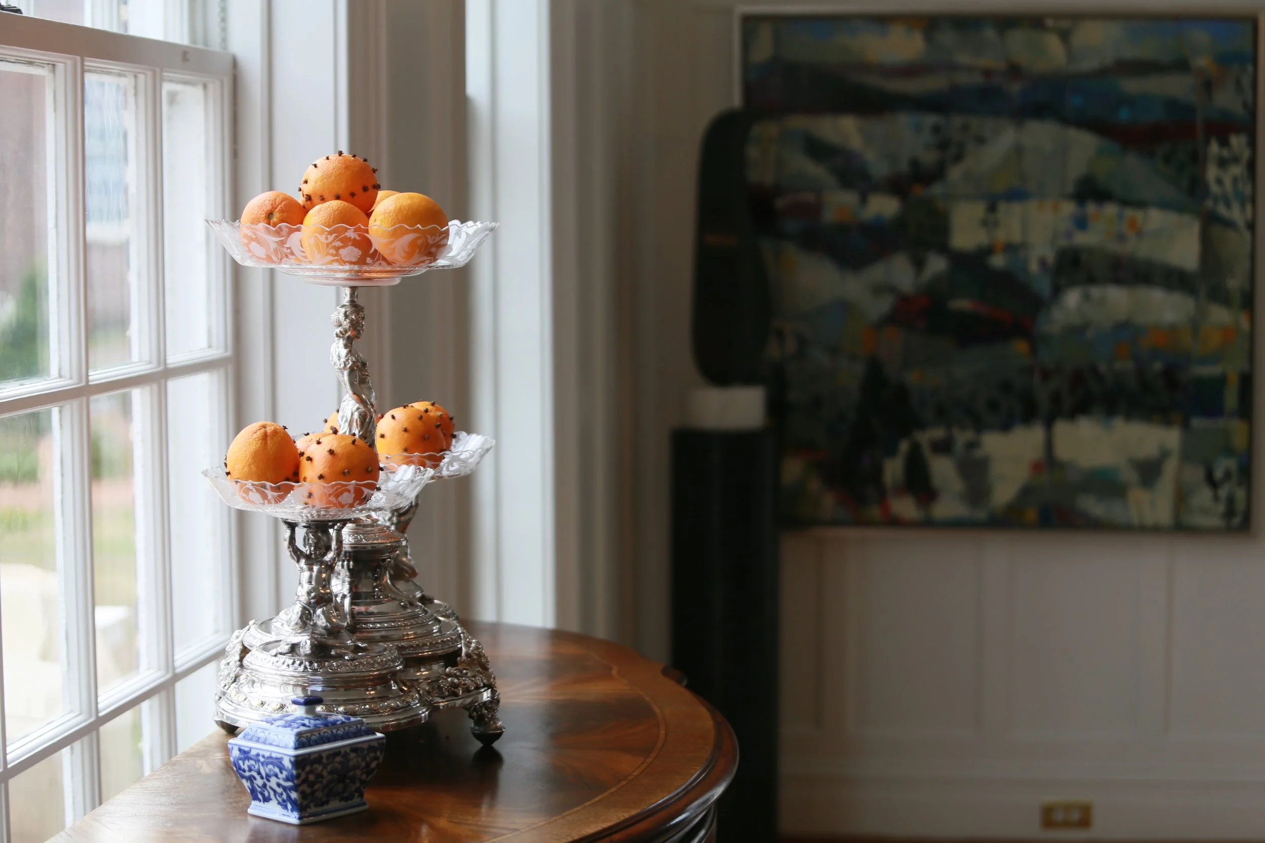 Decorative silver tiered stand with orange pastries topped with black sprinkles, set on a wooden table next to a window in a room with white paneling and colorful abstract art on the wall.
