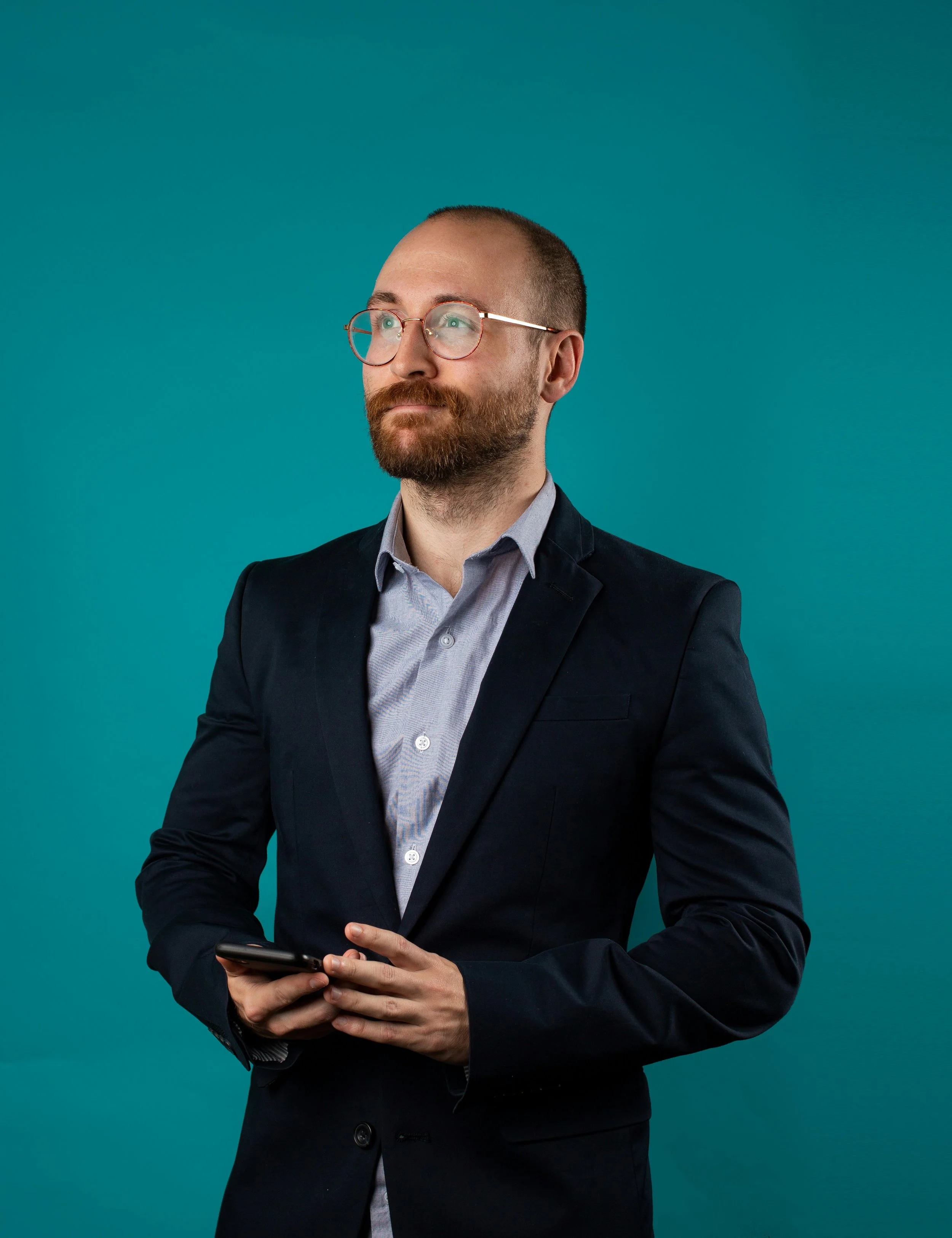 A man in a business suit with glasses is looking slightly to his right while holding a smartphone in his hands against a teal background.