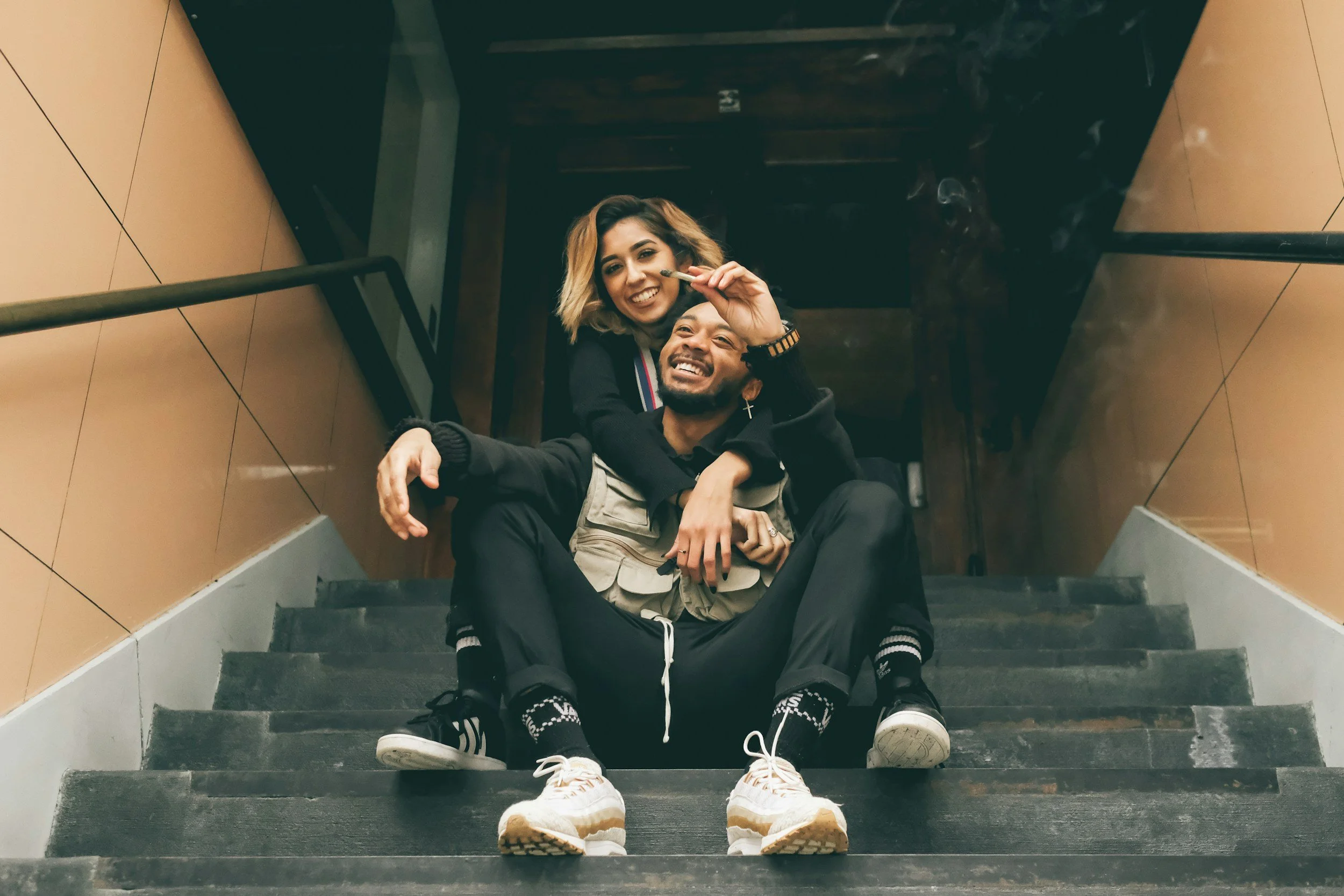 Two people sitting on stairs, one man and one woman, smiling, with the woman holding a lit cigarette, in a relaxed and happy pose.