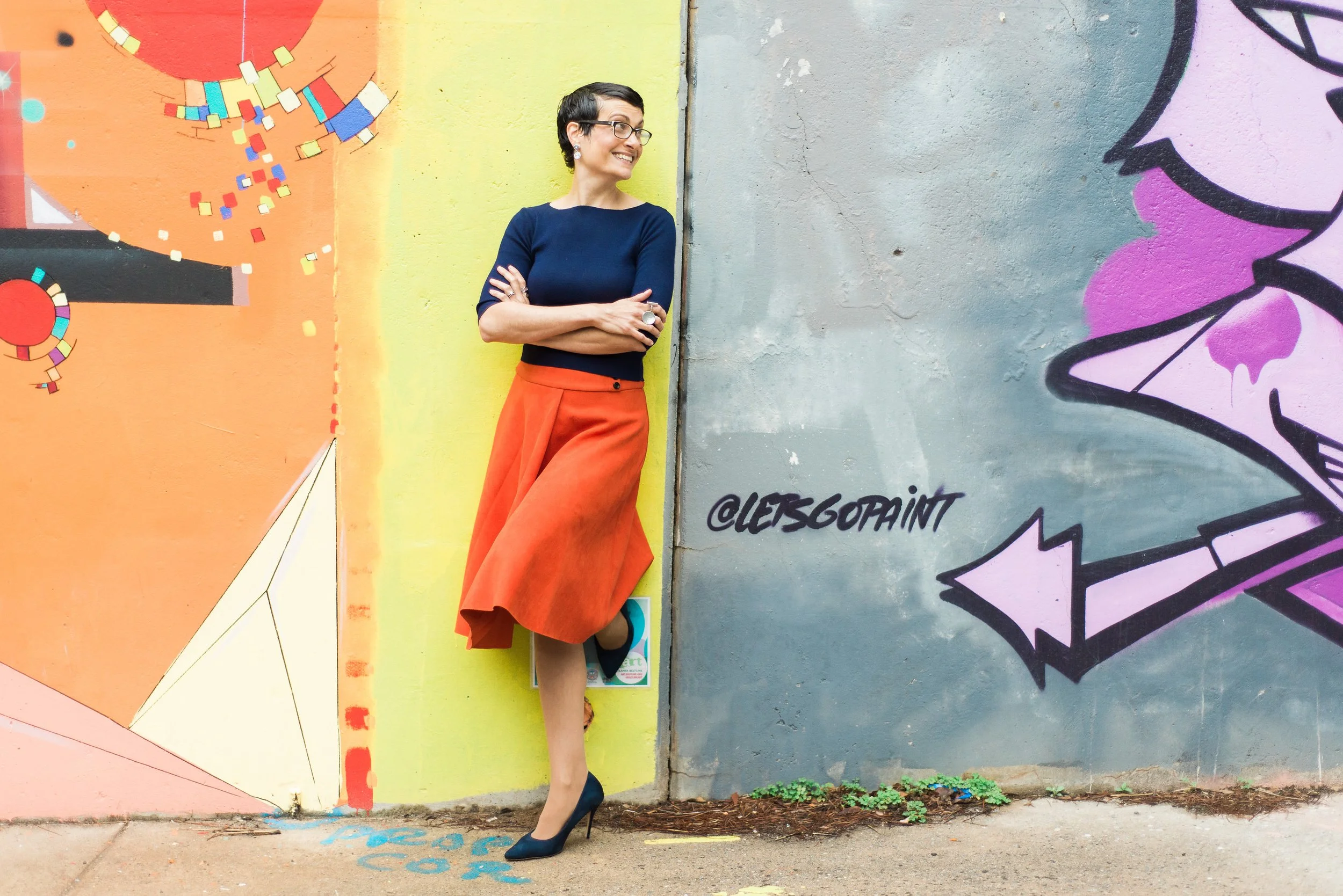 Woman standing against a colorful graffiti wall, smiling, wearing a navy top, orange skirt, black heels, glasses, and earrings.