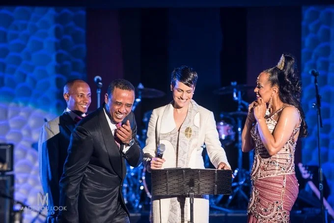 Dawn with big smile in cream suit with stole standing at a music stand with a groom on one side in a suit with a purple tie laughing and a bried on the other side smiling in a purple and gold outfit