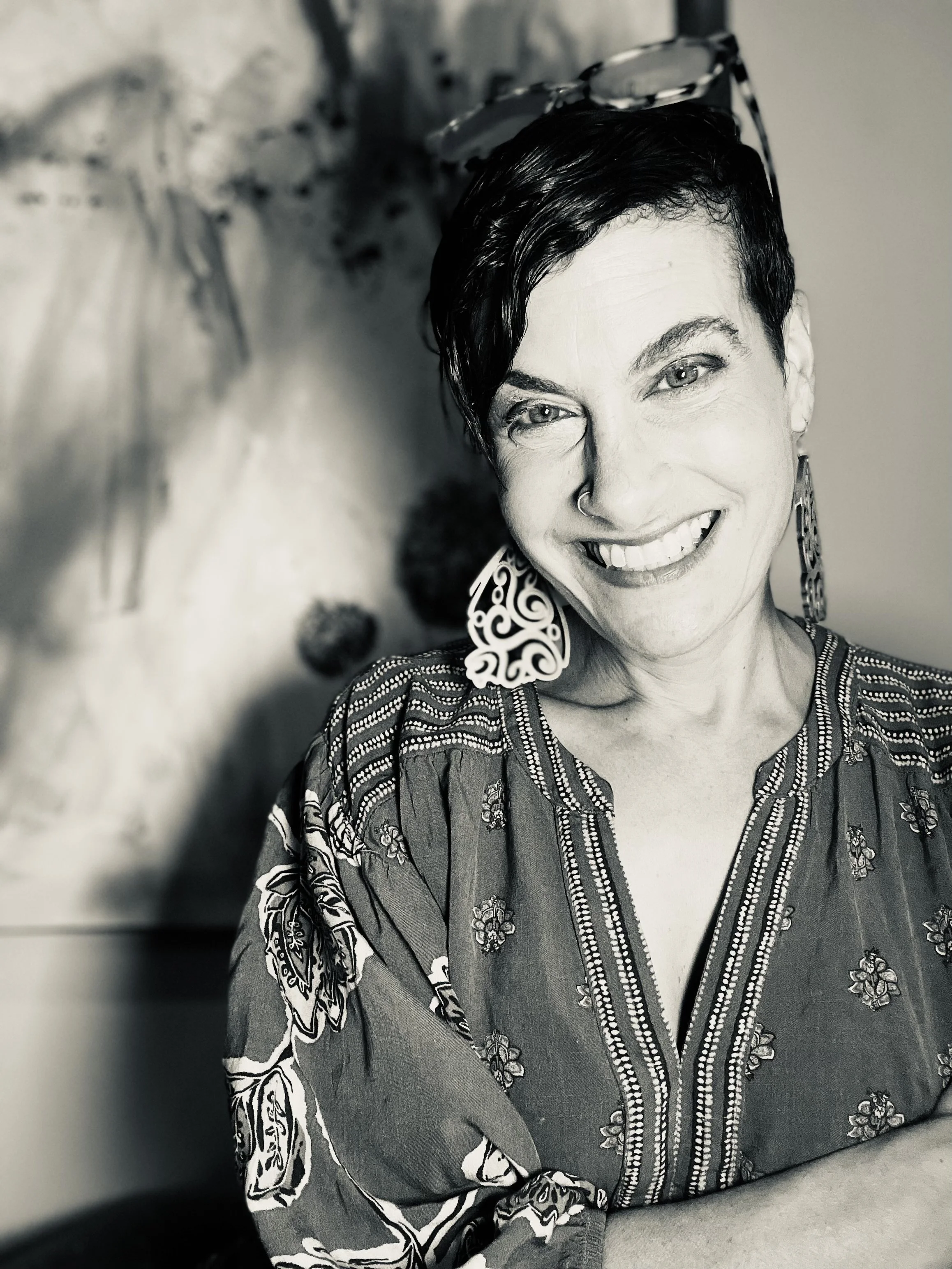 A black and white photo of a smiling woman with short dark hair, wearing earrings and a patterned top, with glasses resting on her head.