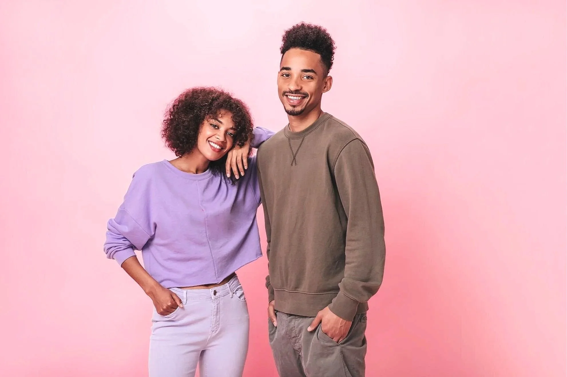 A young woman and man standing close together, smiling, against a pink background. The woman has curly dark hair and is wearing a purple sweatshirt and white jeans. The man has short curly hair and is wearing a brown sweatshirt and gray pants.