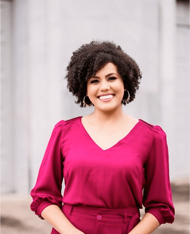 Smiling woman in magenta top and matching pants outdoors, with curly hair and hoop earrings.