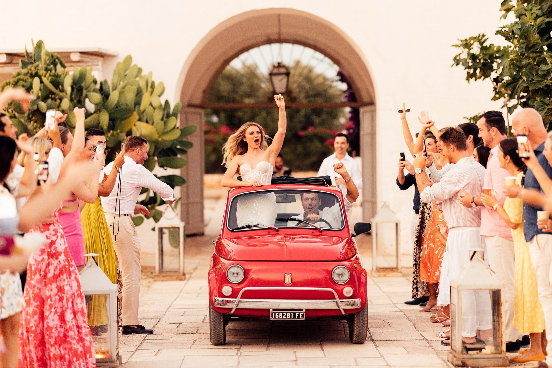 bride raises hand as couple enter wedding venue in vintage red fiat at masseria potenti