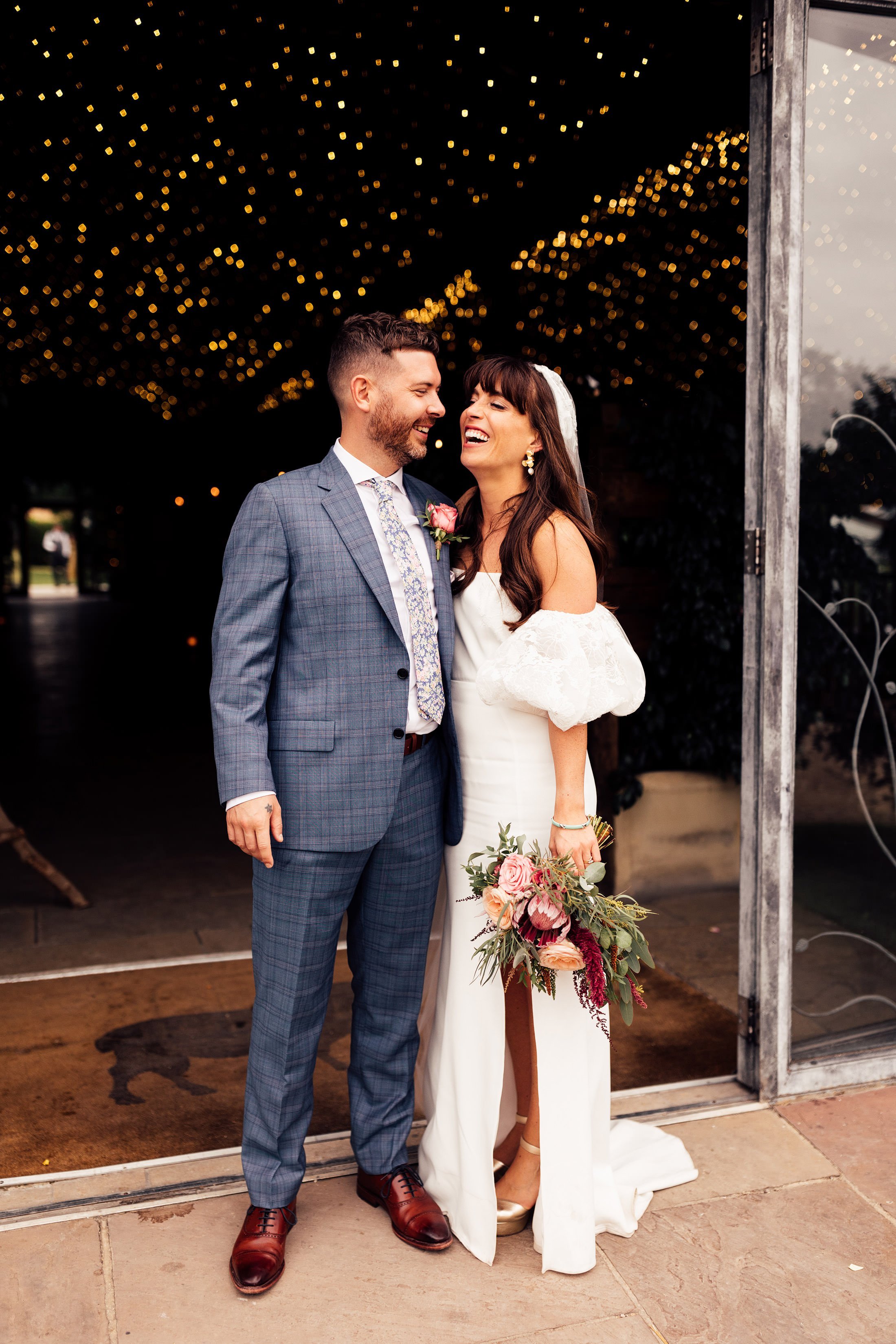 bride and groom laugh and embrace as bride holds colourful floral bouquet  and wears rime Arodaky dress complete with Miu miu shoes