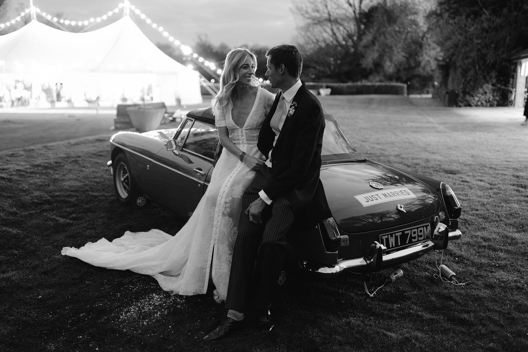 black and white photo of bride and groom posing on vintage car with marquee in the background