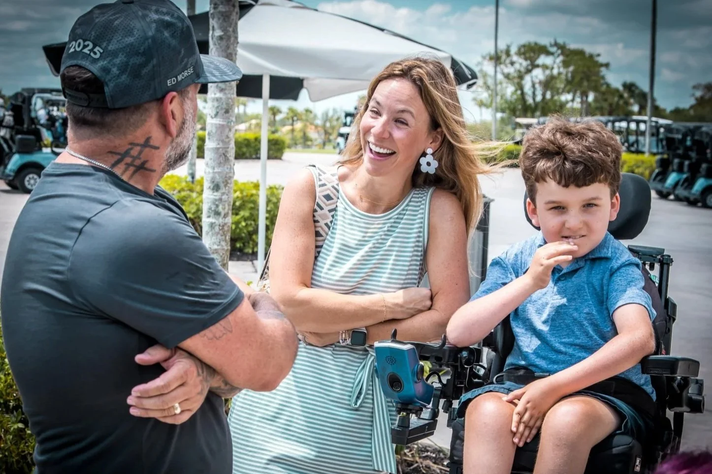 A woman smiling and talking to a man with tattoos on his arms and neck, and a boy in a wheelchair, outdoors near a parking lot with golf carts and trees.