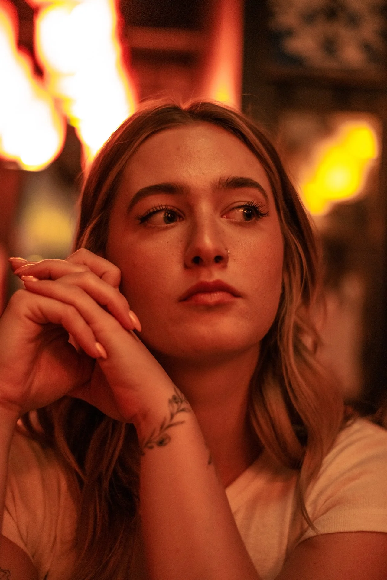 Close-up of a young woman with long wavy hair, resting her chin on her hands, in a dimly lit setting with orange and yellow lights in the background.