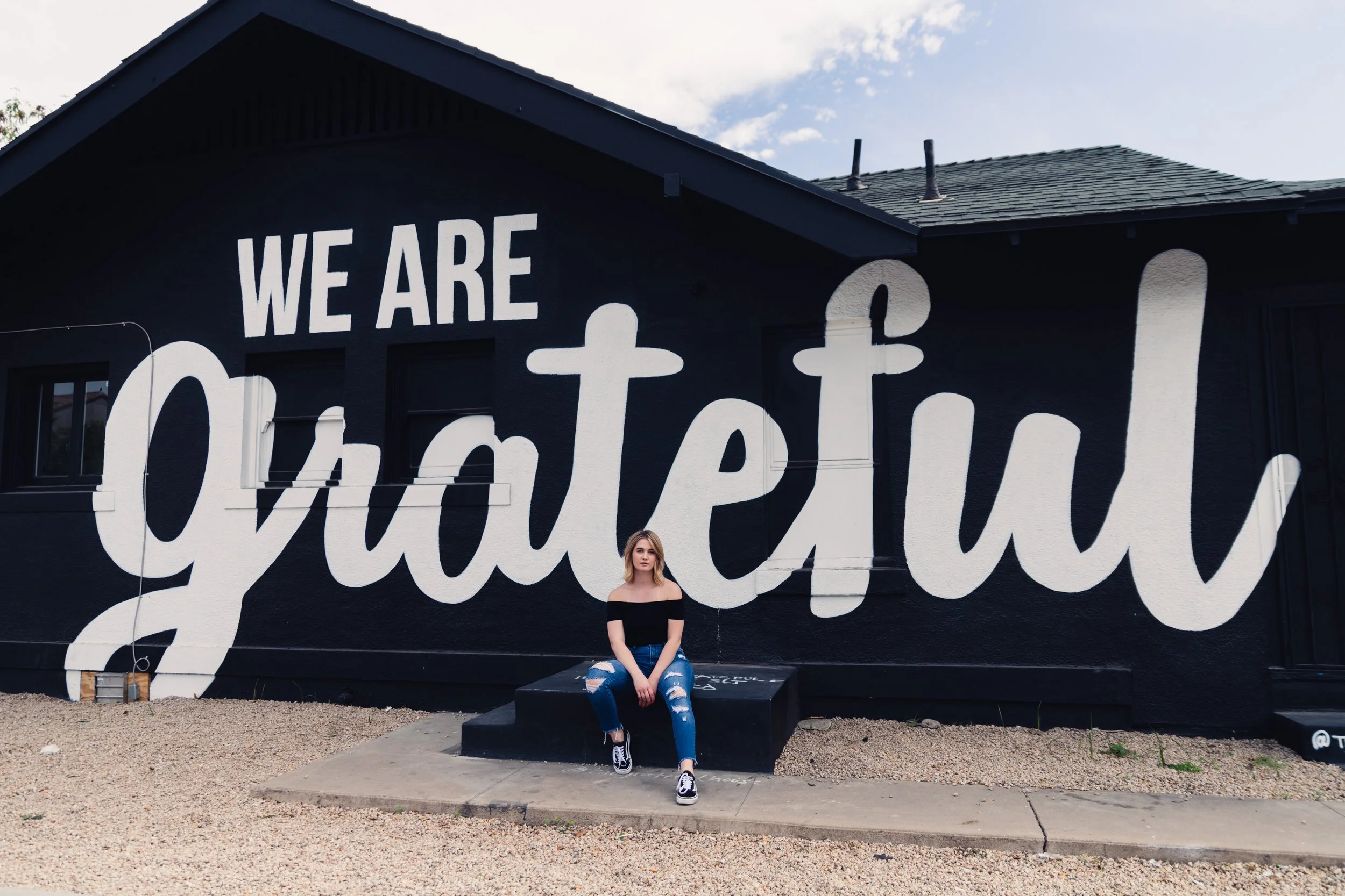 A person sitting on a black platform in front of a large mural that reads 'We are grateful' in white letters on a dark building.