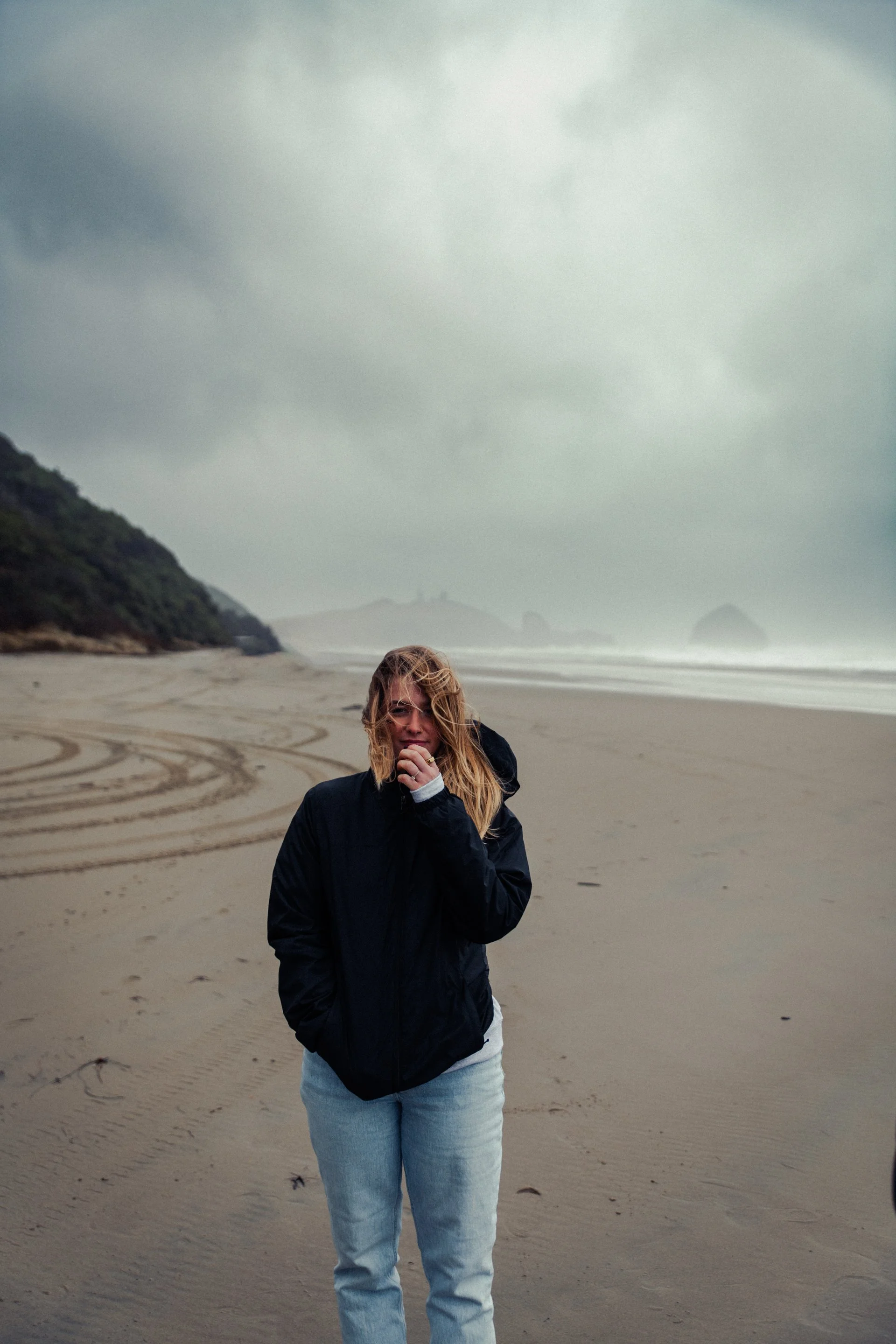 A woman standing on a cloudy beach in a black jacket and light jeans, with ocean waves and rocky islands in the background.