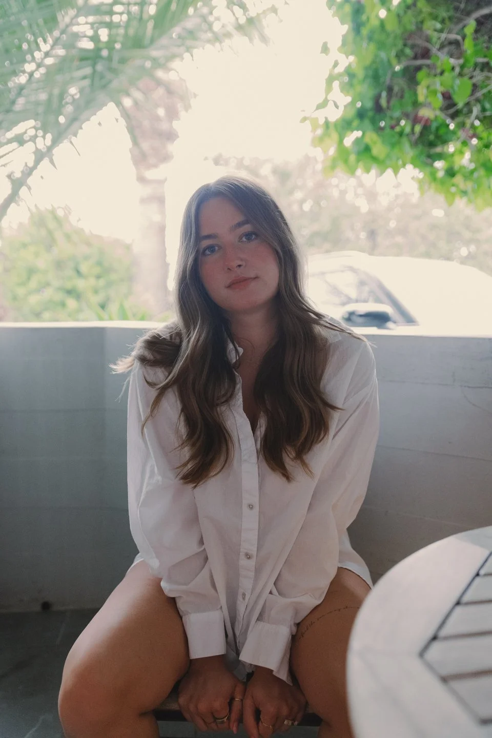 A young woman with long wavy brown hair sitting outdoors in front of a bright window, wearing a loose white button-up shirt, with a relaxed expression.