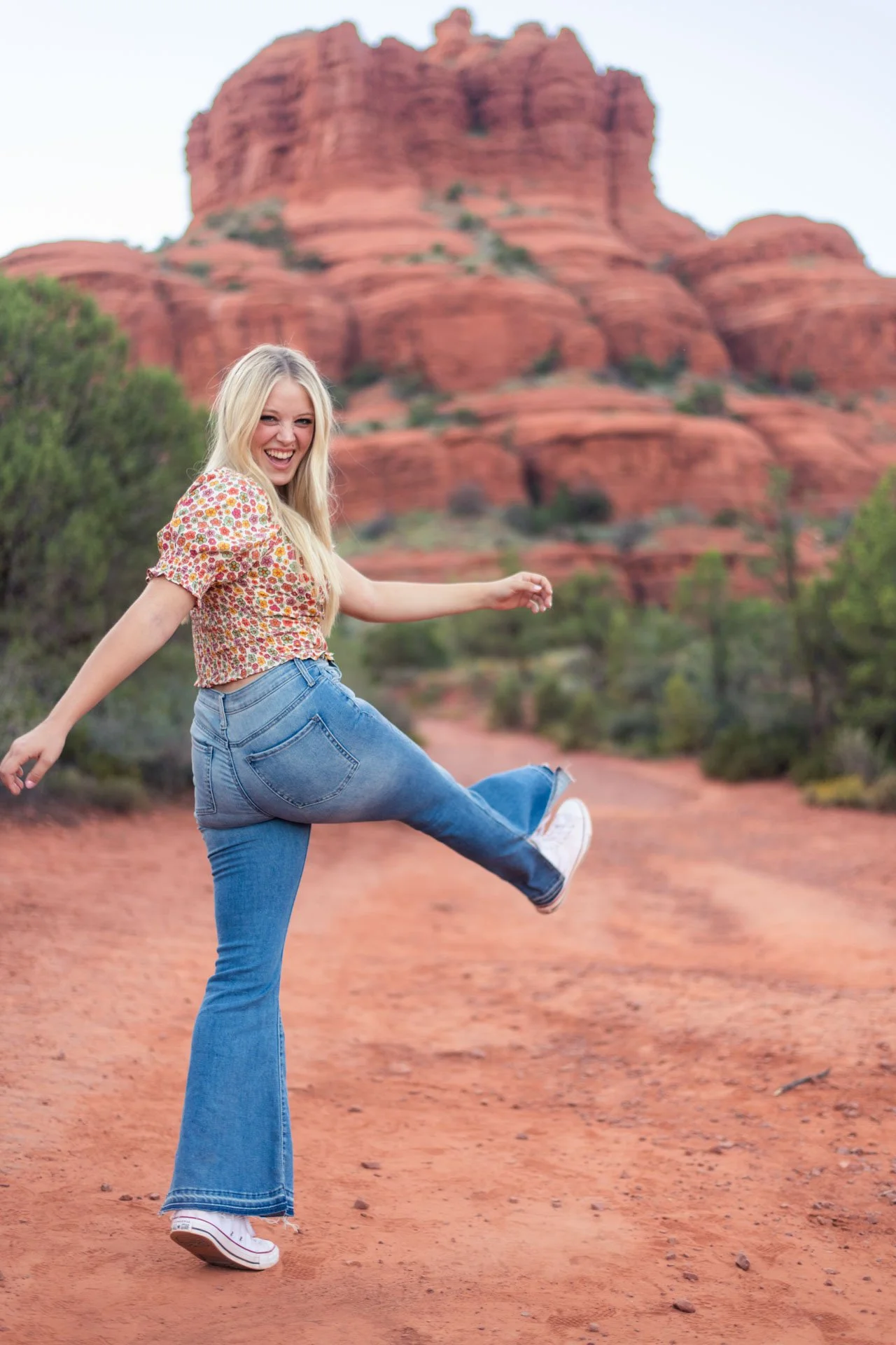 A young woman smiling and kicking her leg in the air on a dirt trail with red rock formations and desert vegetation in the background.