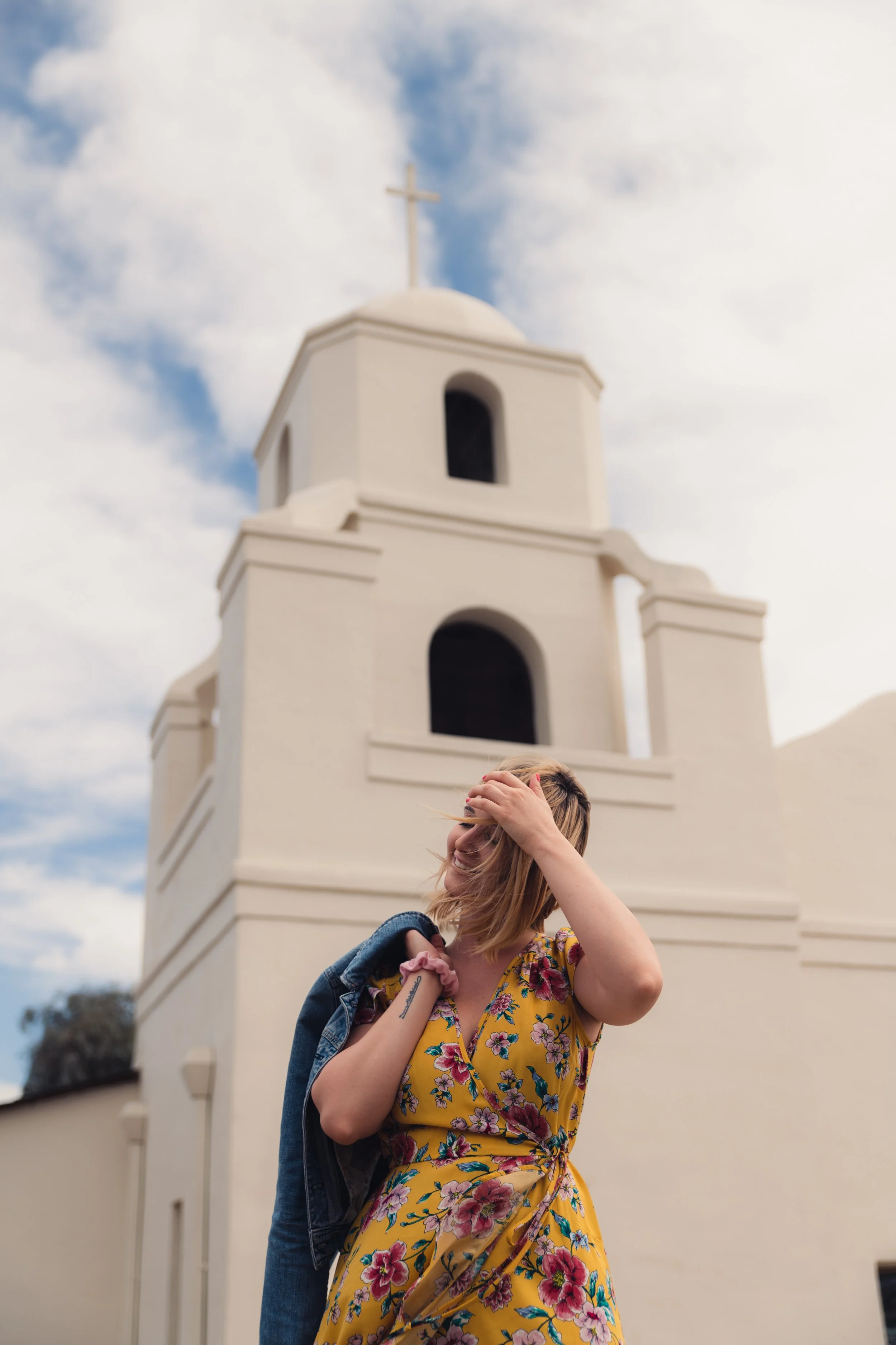 A woman in a yellow floral dress stands outdoors in front of a white church with a cross on top. She is smiling and holding her hair back with one hand, carrying a denim jacket on her shoulder, and the sky is partly cloudy.