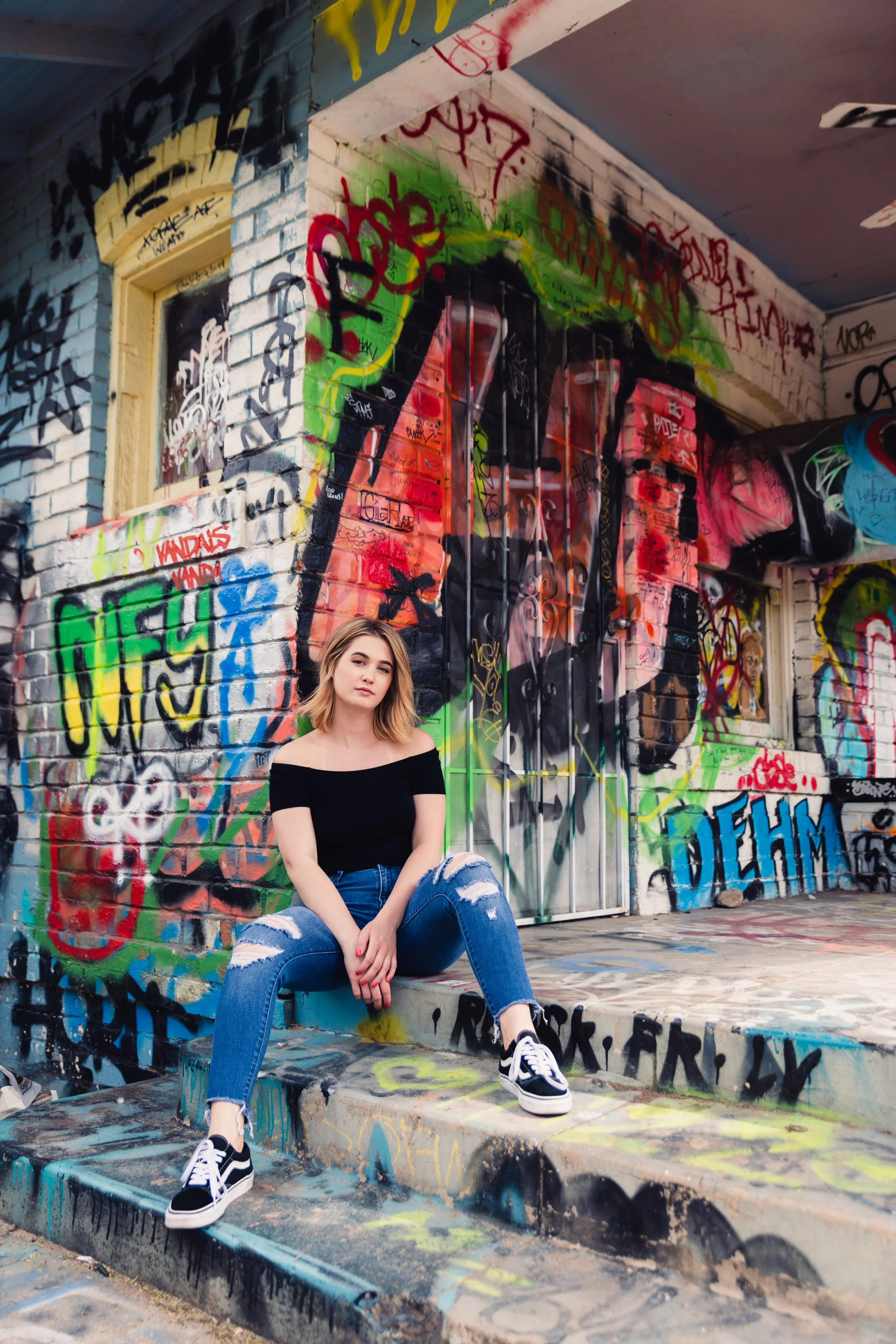 Young woman with blonde hair in a black off-the-shoulder top and ripped jeans sitting on steps covered in graffiti street art, in front of a wall with a large painted face and various tags and colors.
