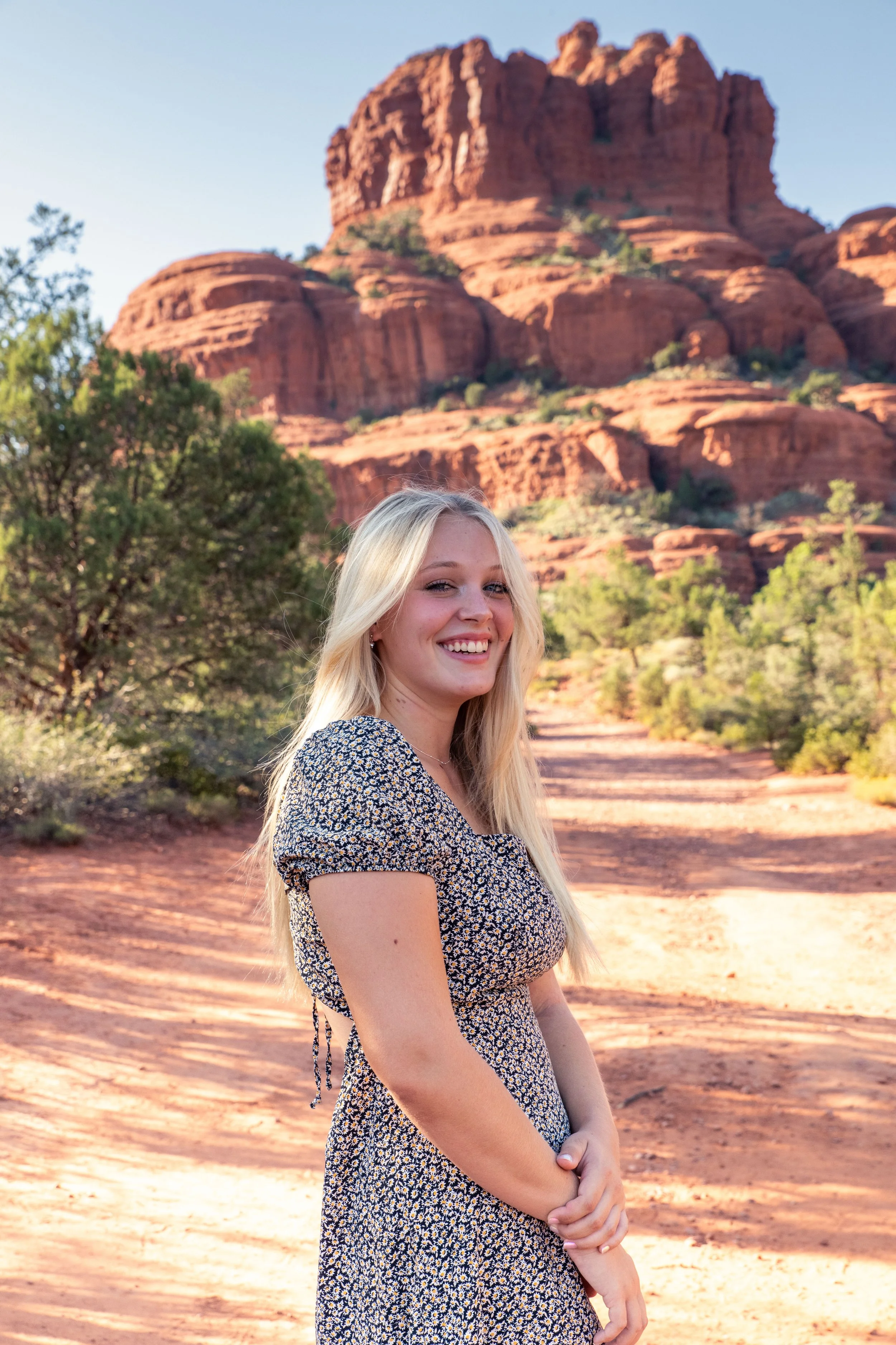 Young woman smiling in desert landscape with red rock formations and mountains in the background.