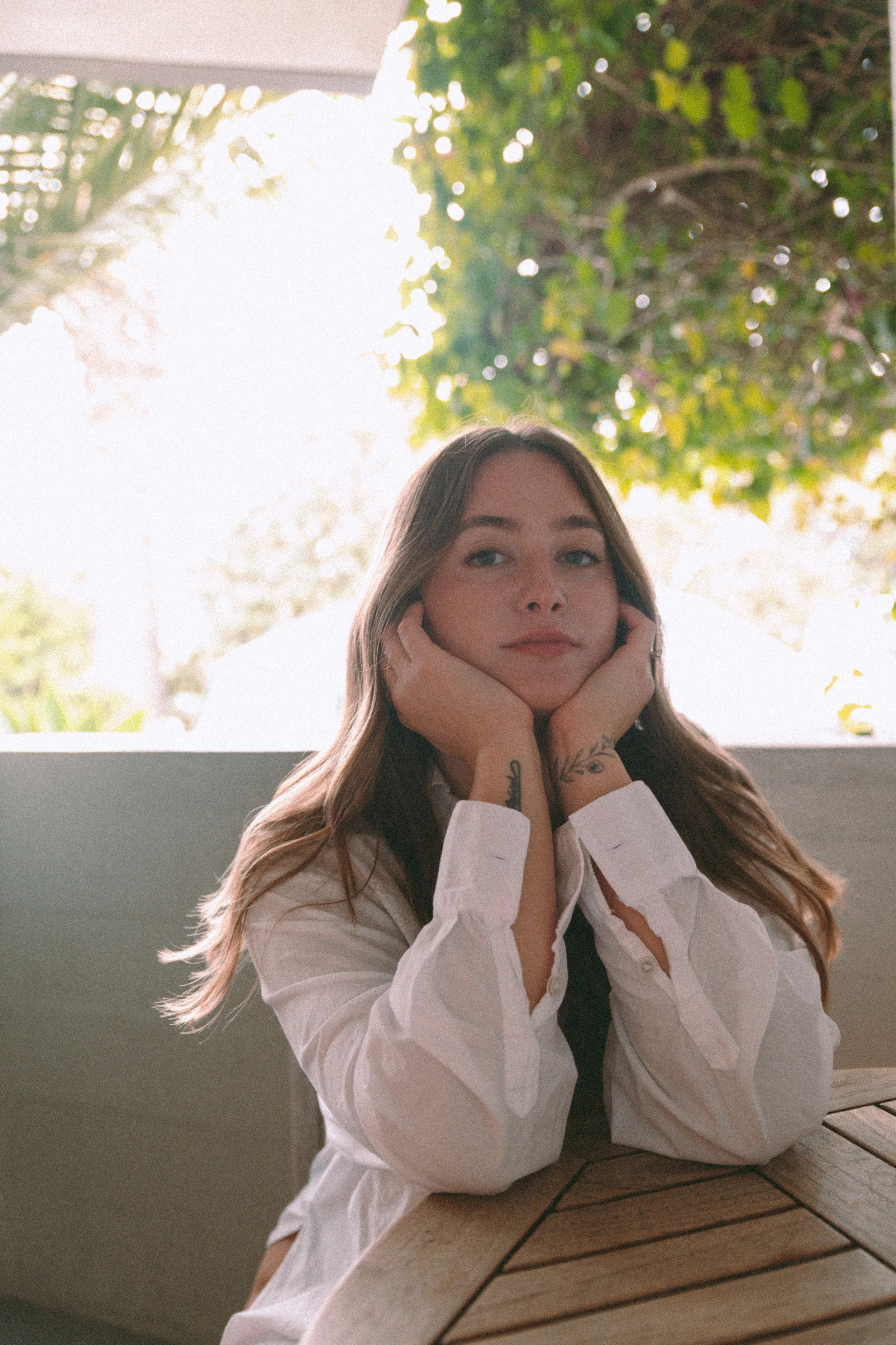 A young woman with light brown hair, wearing a white blouse, sitting at a wooden table outdoors, resting her chin on her hands, with greenery and sunlight in the background.