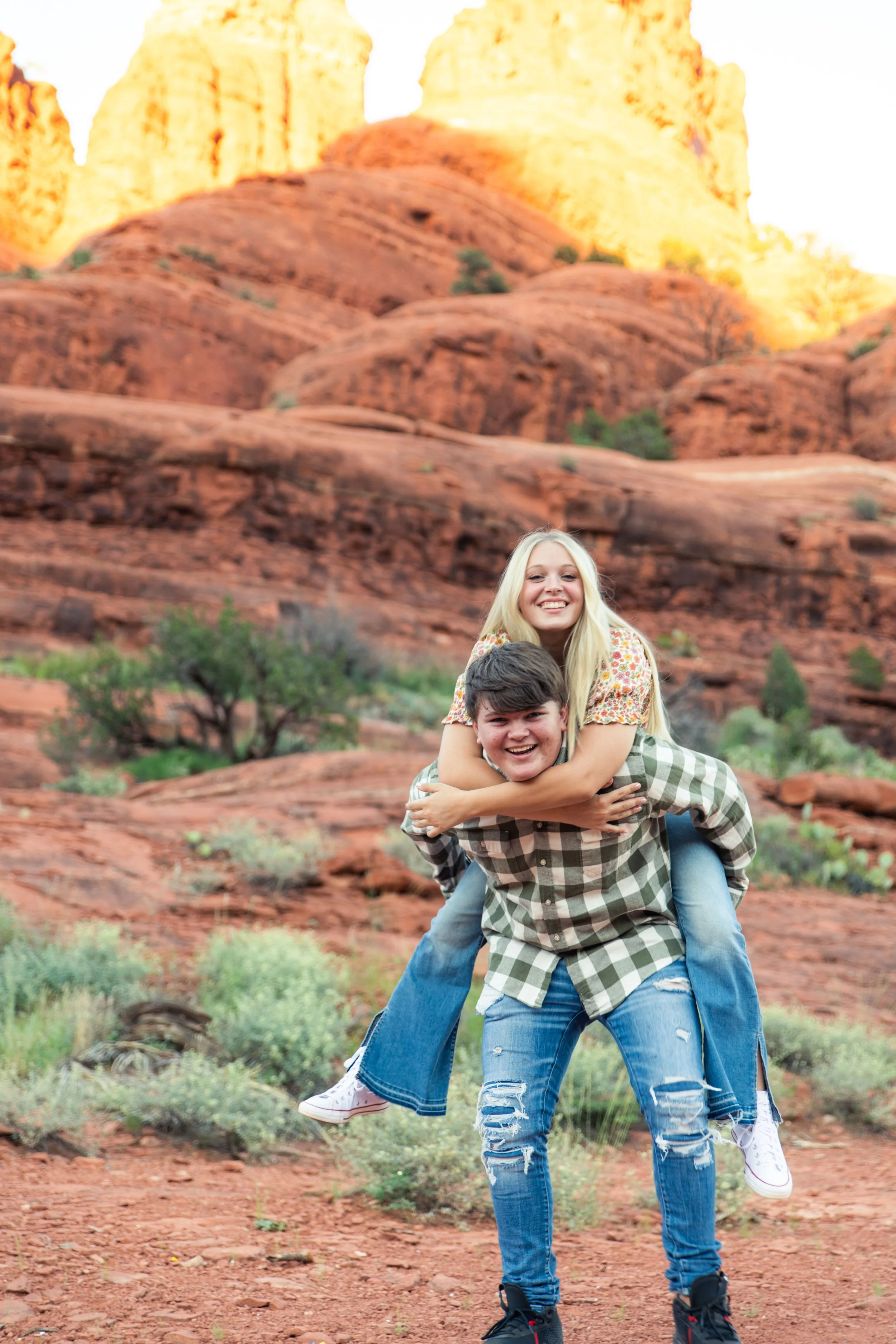 Two teenagers playfully smiling, one giving the other a piggyback ride in a desert landscape with red rocks and greenery, with cliffs in the background.