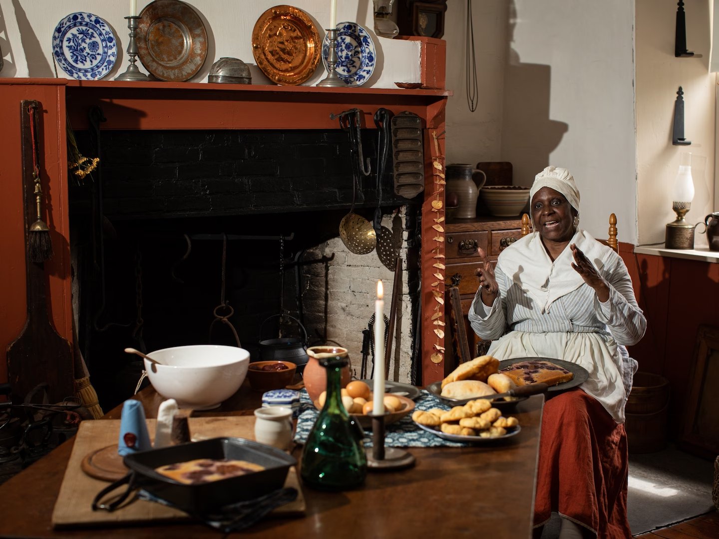 Estelle T Barada, an 18th century historic re-enactor, in costume as Duchess Quamino. Barada is seated amongst a kitchen setting set to the late 18th century behind a table with food and dishes with a fireplace in the background.