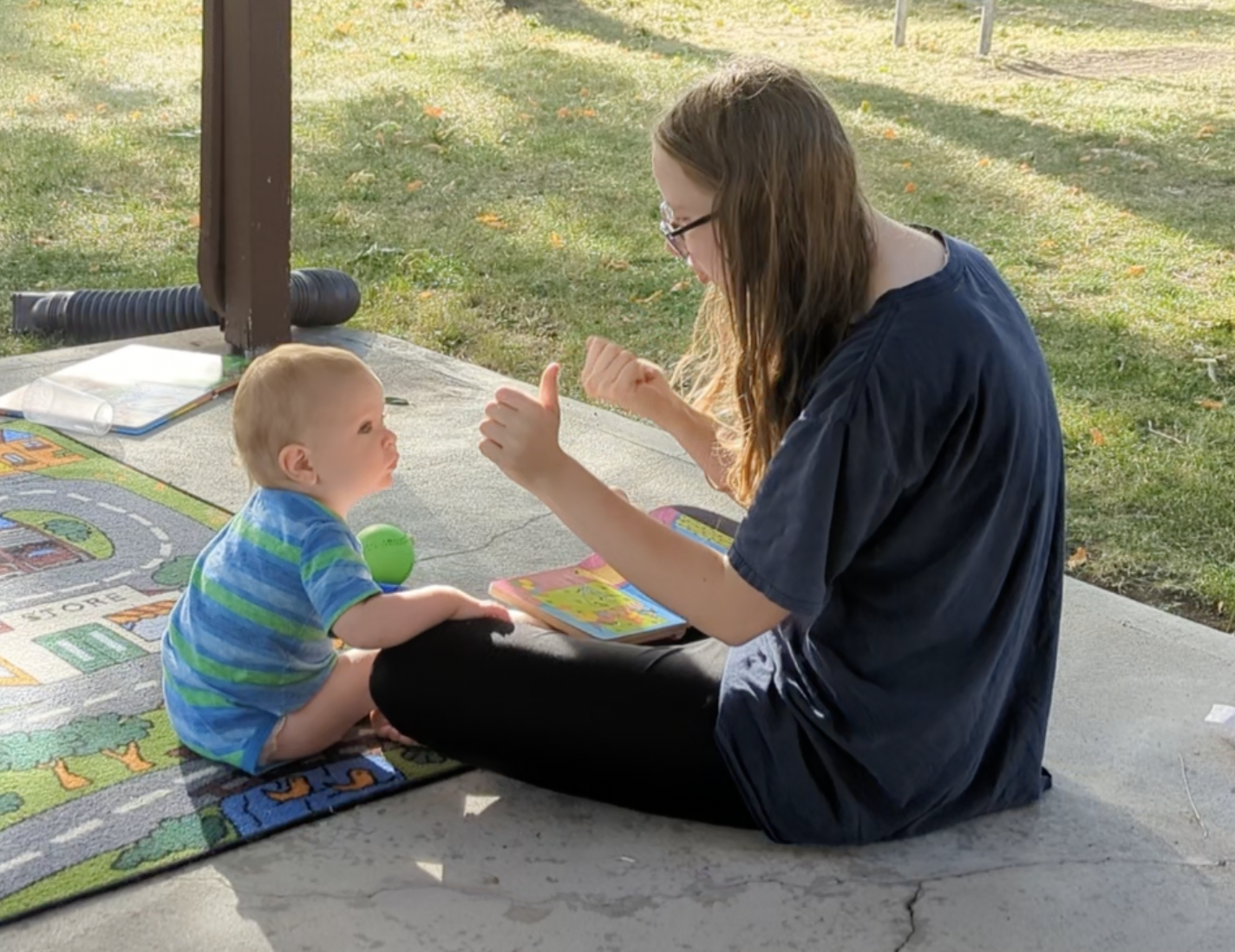 toddler and young woman, both white, sitting outside and looking at a picture book